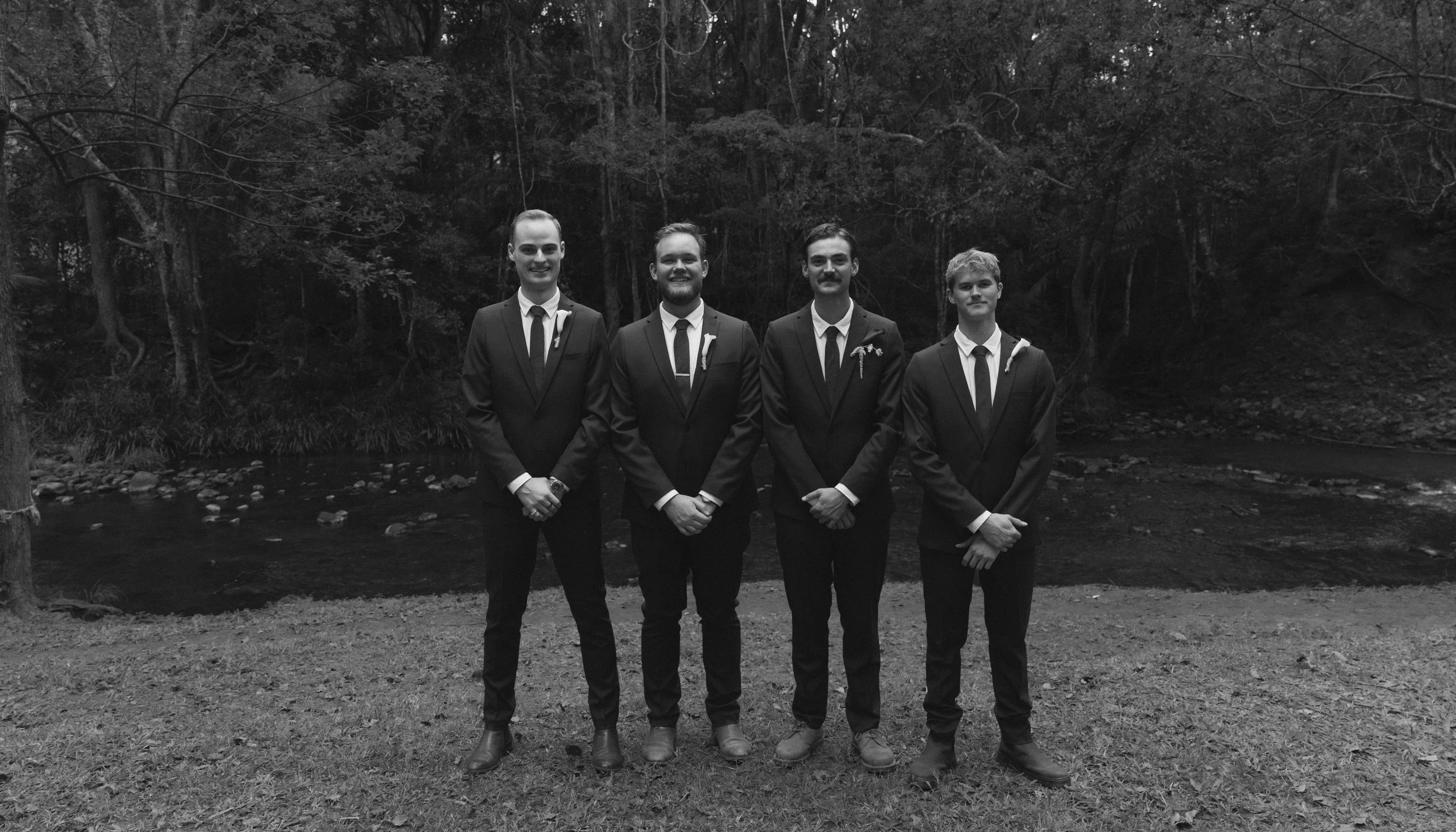 Four men dressed in formal suits and ties standing outdoors in front of a river and wooded area.