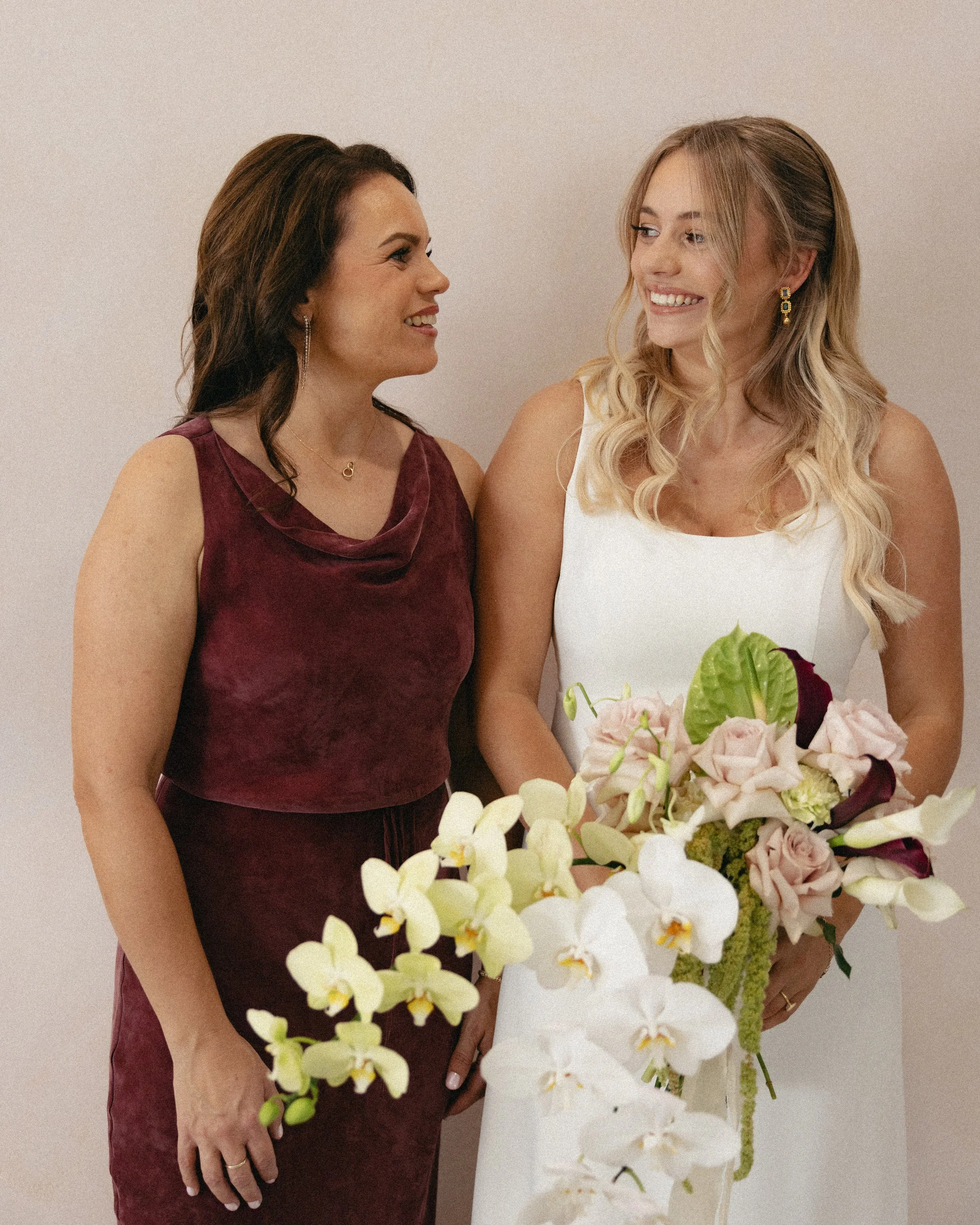 Two women smiling at each other, one holding a large bouquet of pink and white flowers, against a plain white wall.