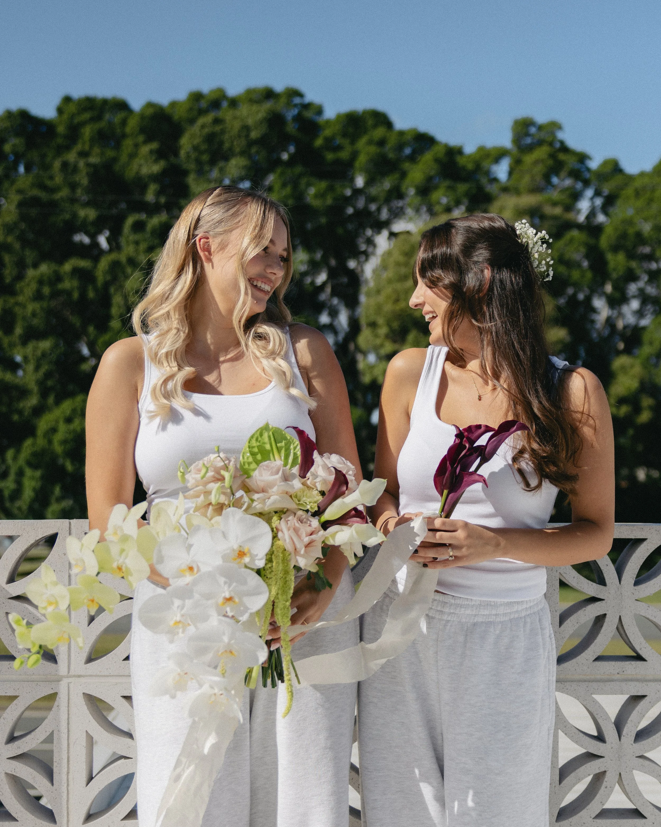 Two women in white tops and casual pants smiling and holding flowers outdoors against a background of trees and a decorative white wall.