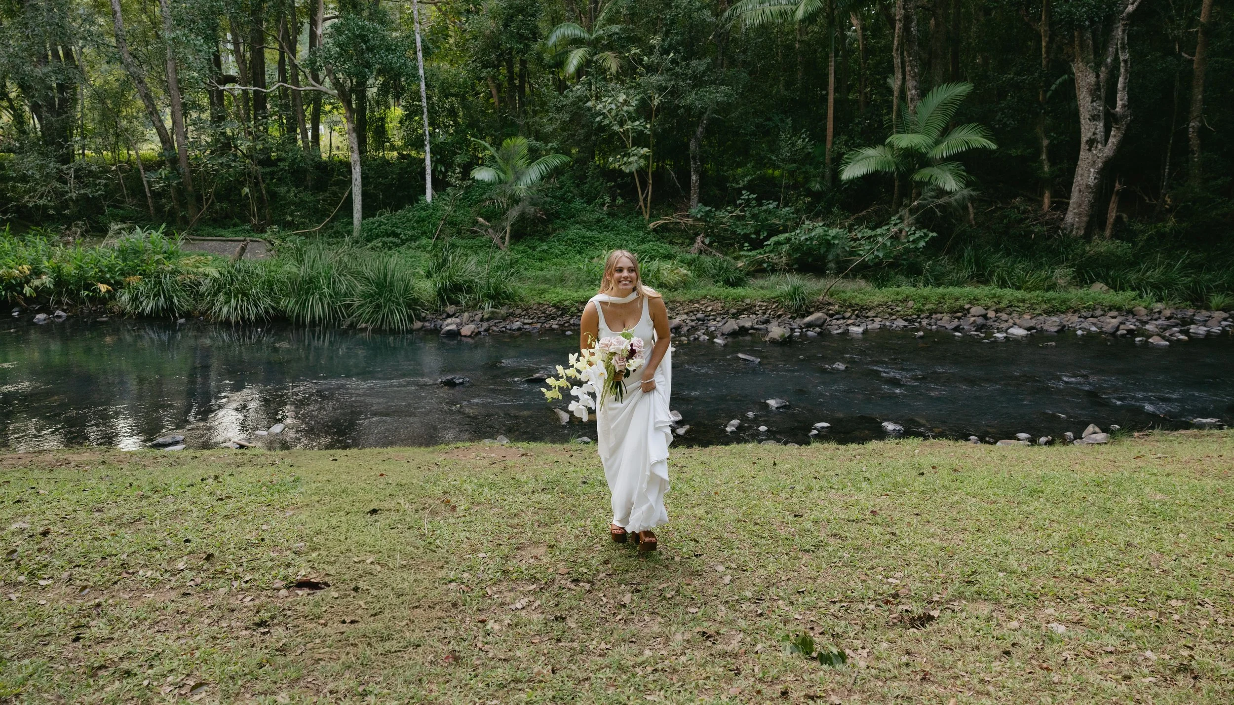 A woman in a white dress holding a bouquet of flowers, standing on grass in front of a river and lush green trees.