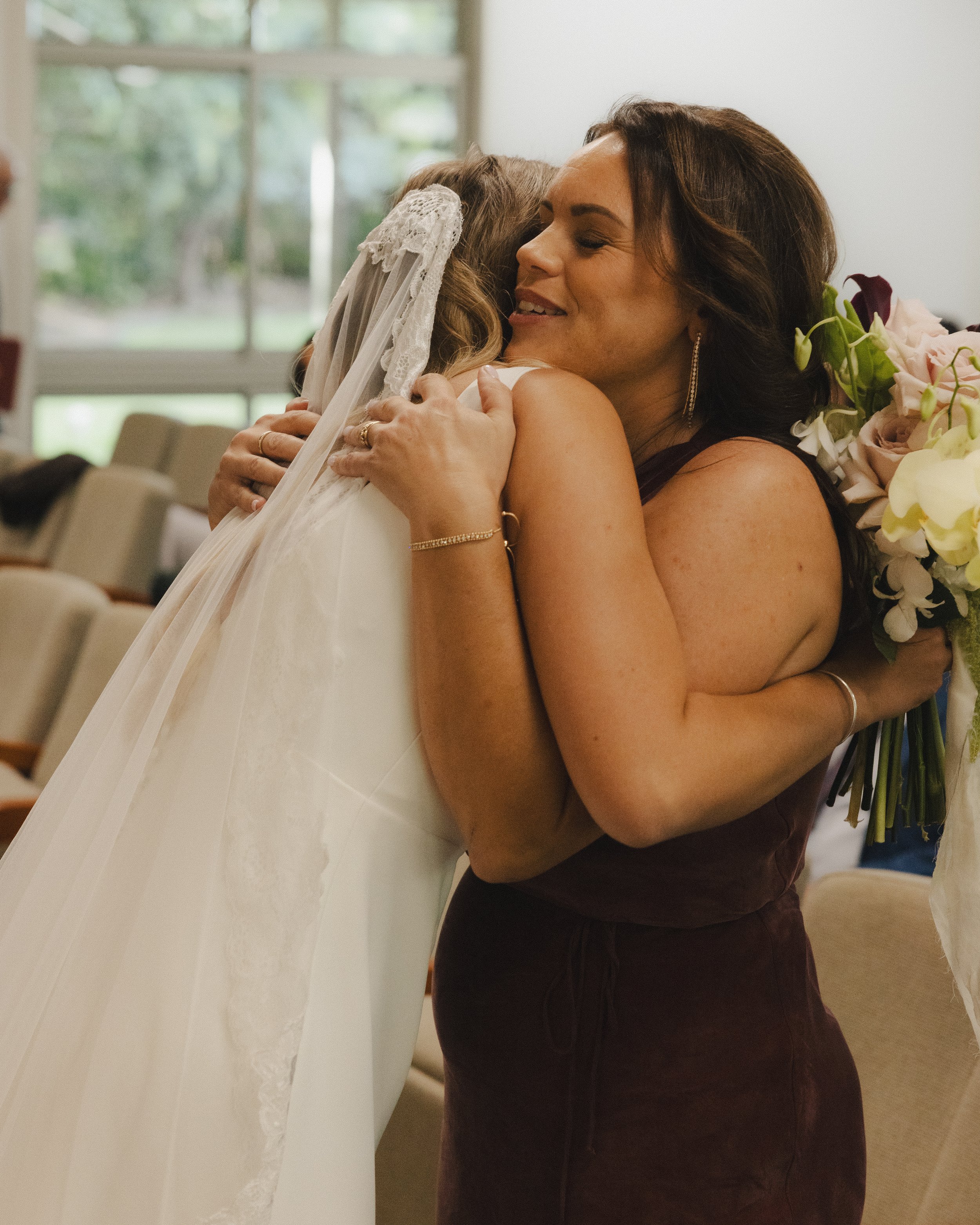 A woman in a dark dress hugging a young girl dressed as a bride, both smiling warmly, in a bright room with large windows and garden view.