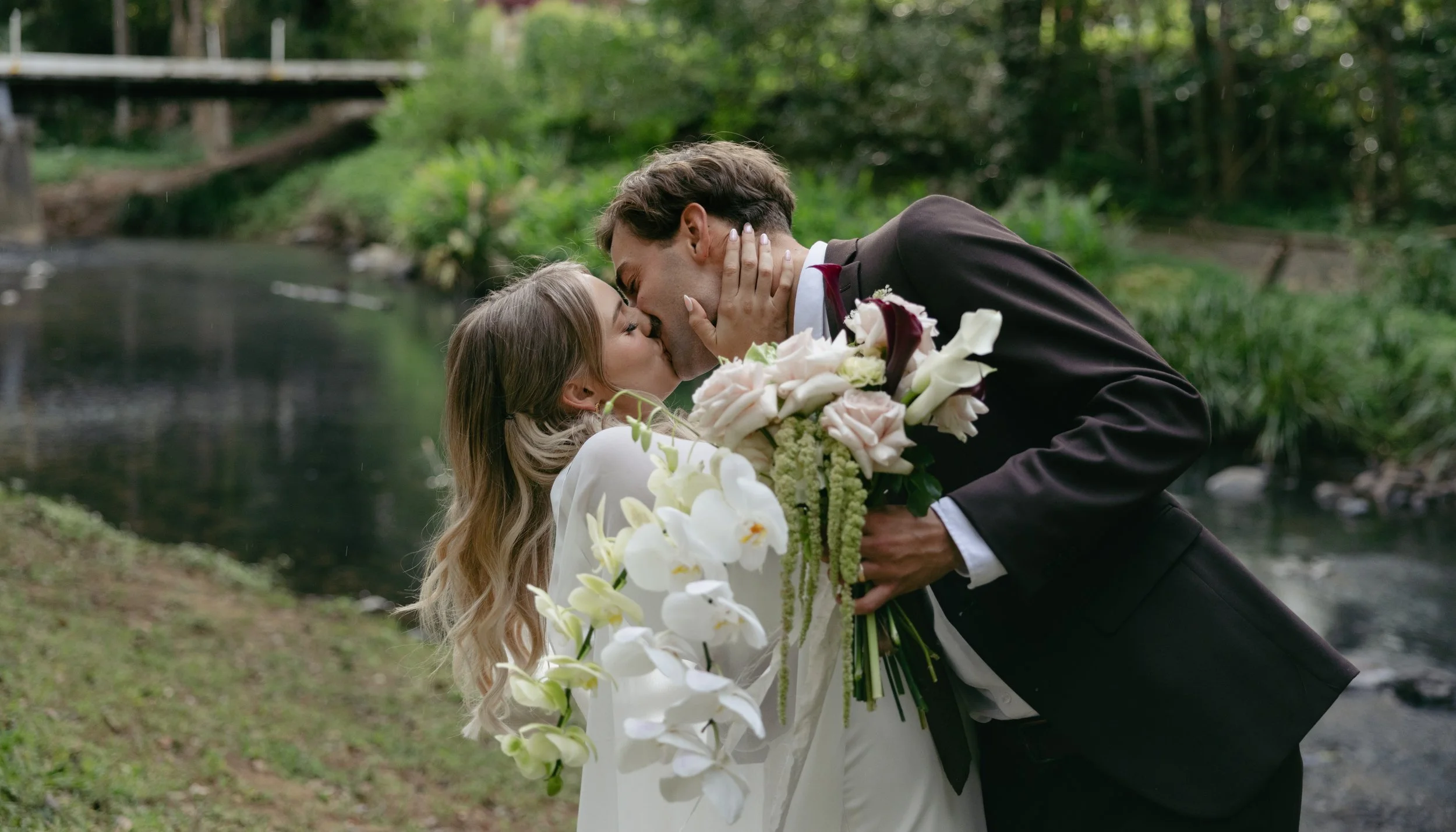 A newlywed couple kisses outdoors near a river, with the groom in a black suit and the bride in a white dress holding a bouquet of flowers.