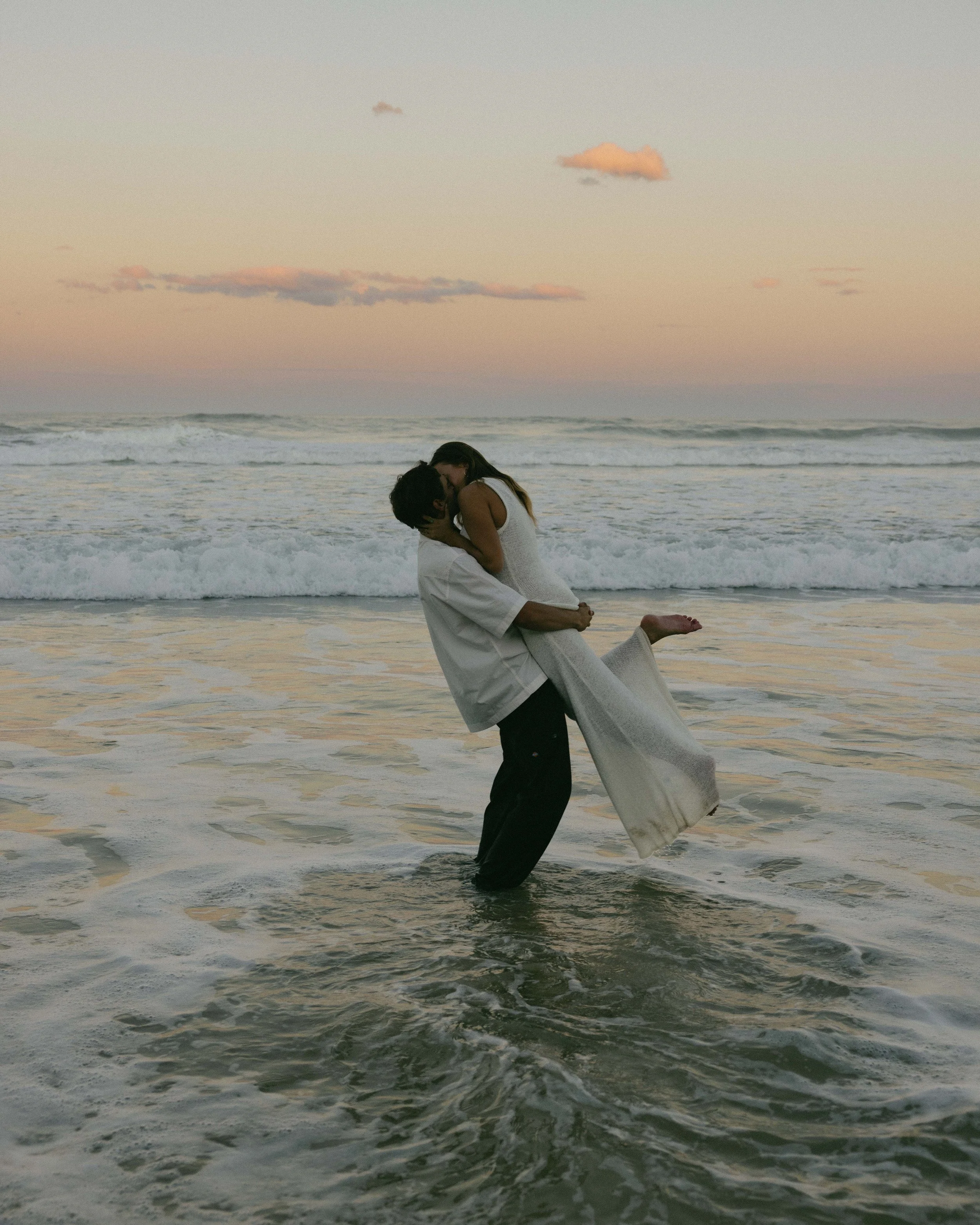 A couple is standing in the ocean at sunset, with the man lifting the woman in his arms. They are dressed in white clothing, and the sky has pastel pink and purple hues.