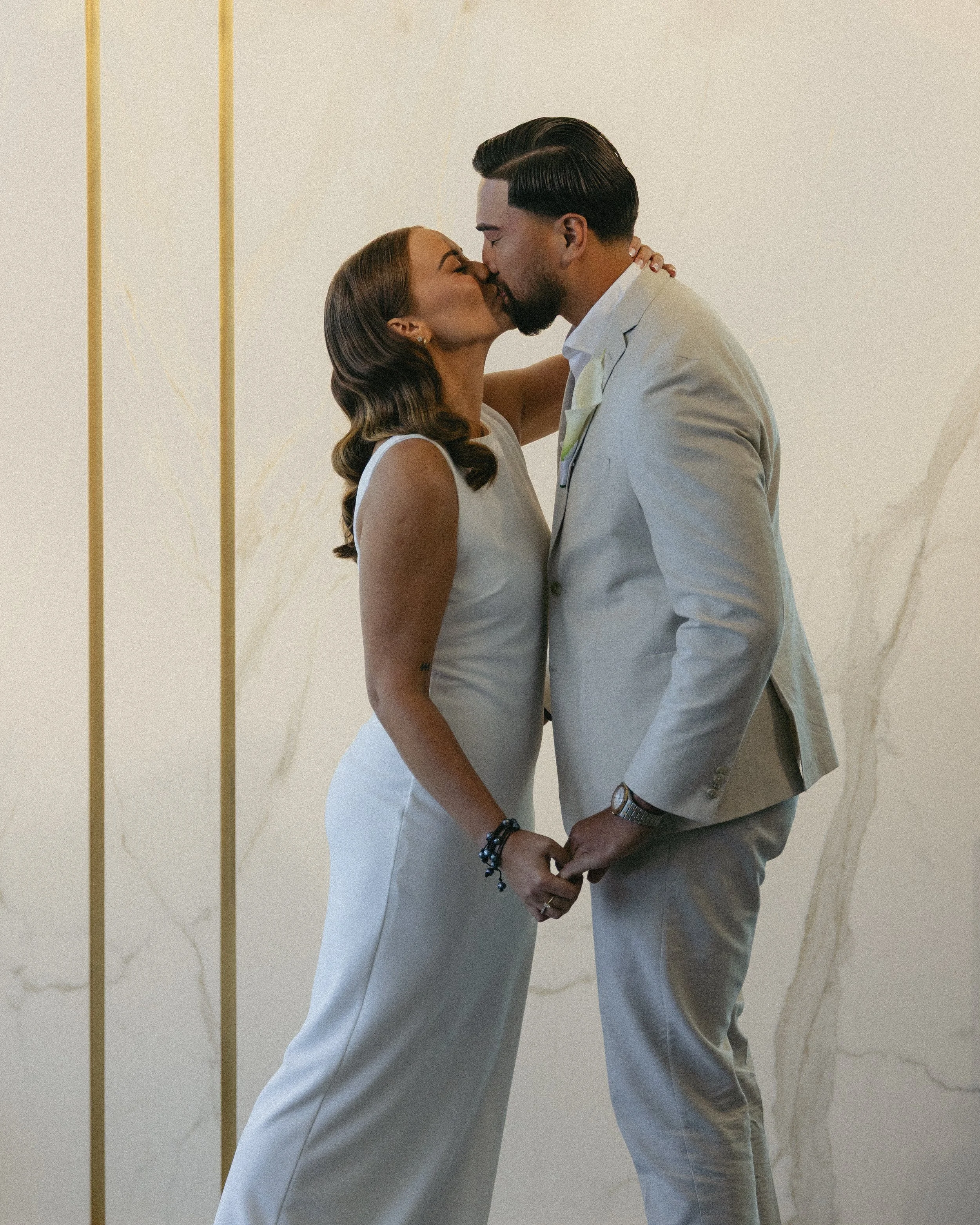 A couple dressed in white, kissing each other, standing hand in hand in front of a marble wall background.