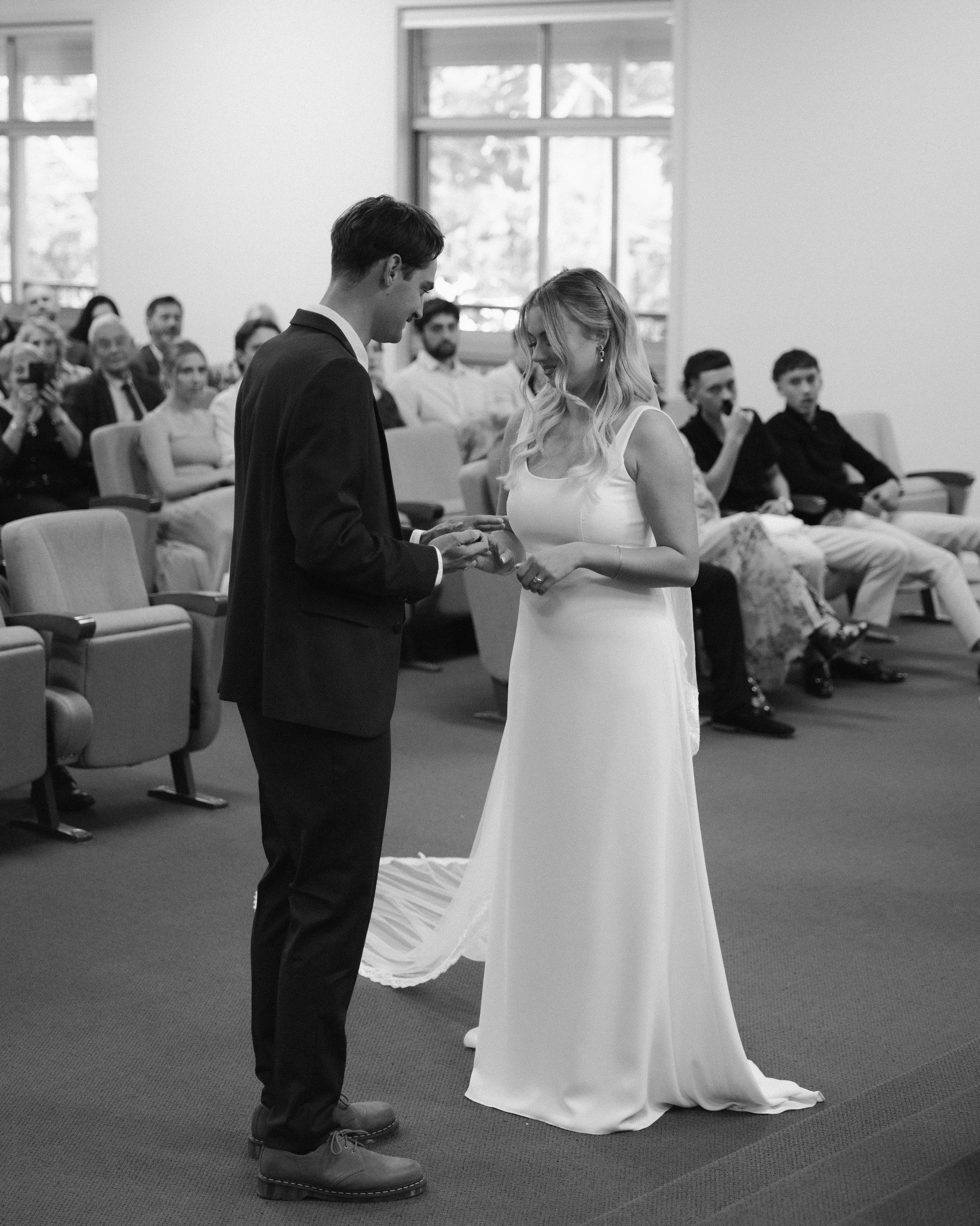 A black and white photo of a wedding ceremony with a bride and groom exchanging rings, surrounded by seated guests in an indoor setting.
