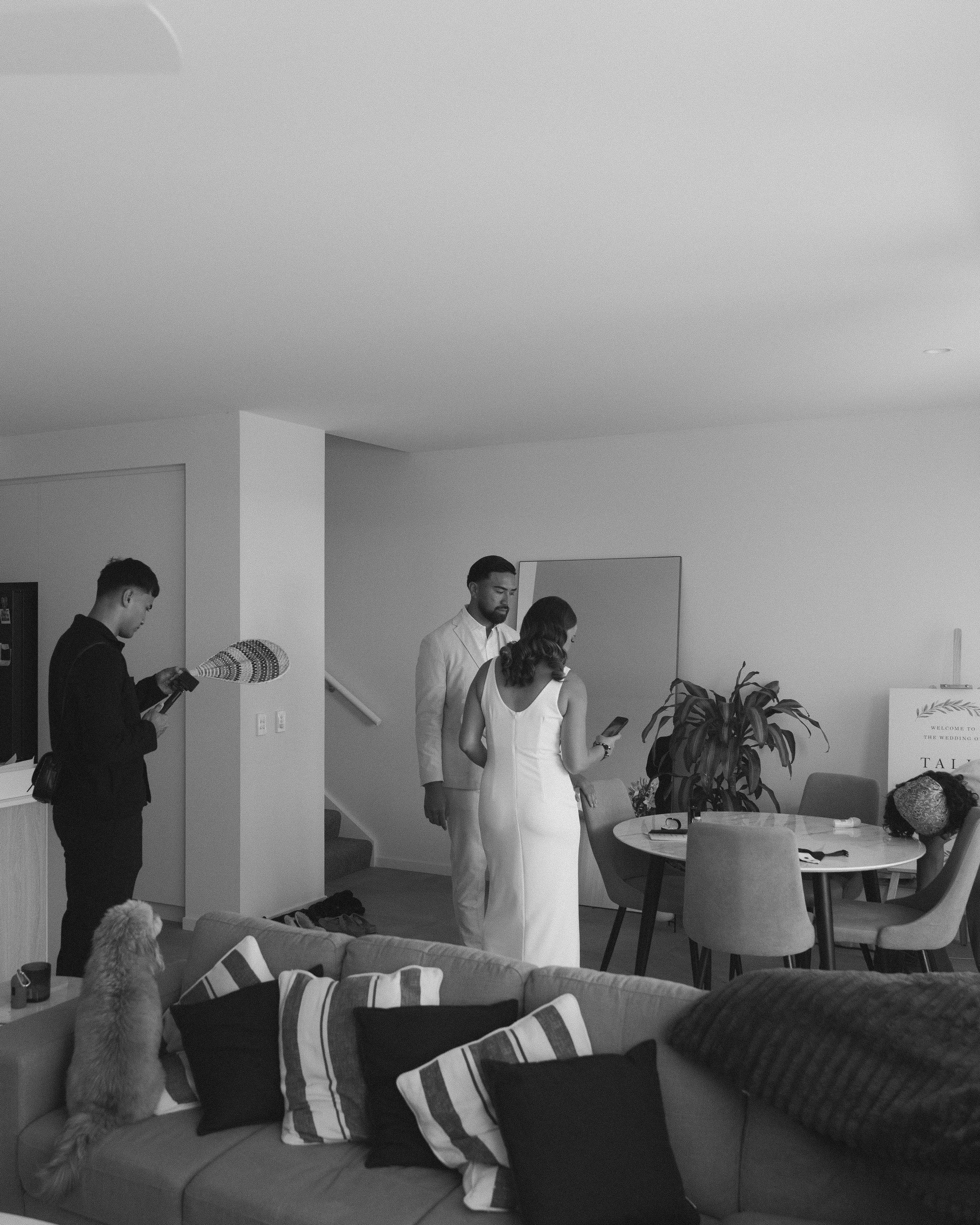A wedding scene indoors with three people, a woman in a white wedding dress, a man in a light suit, and another woman. There is a dog sitting on a couch and a person with a microphone recording or filming. The room has a round table, chairs, and a large potted plant, with some items on the table and a welcome sign.
