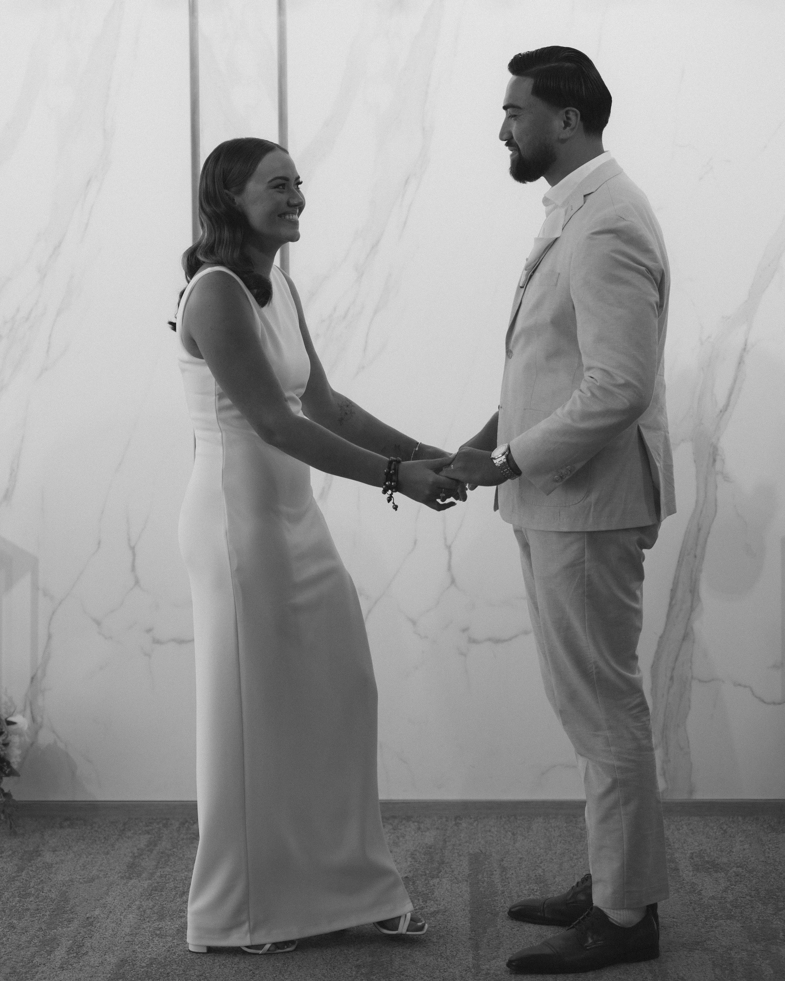A man and woman in wedding attire holding hands and smiling at each other indoors.