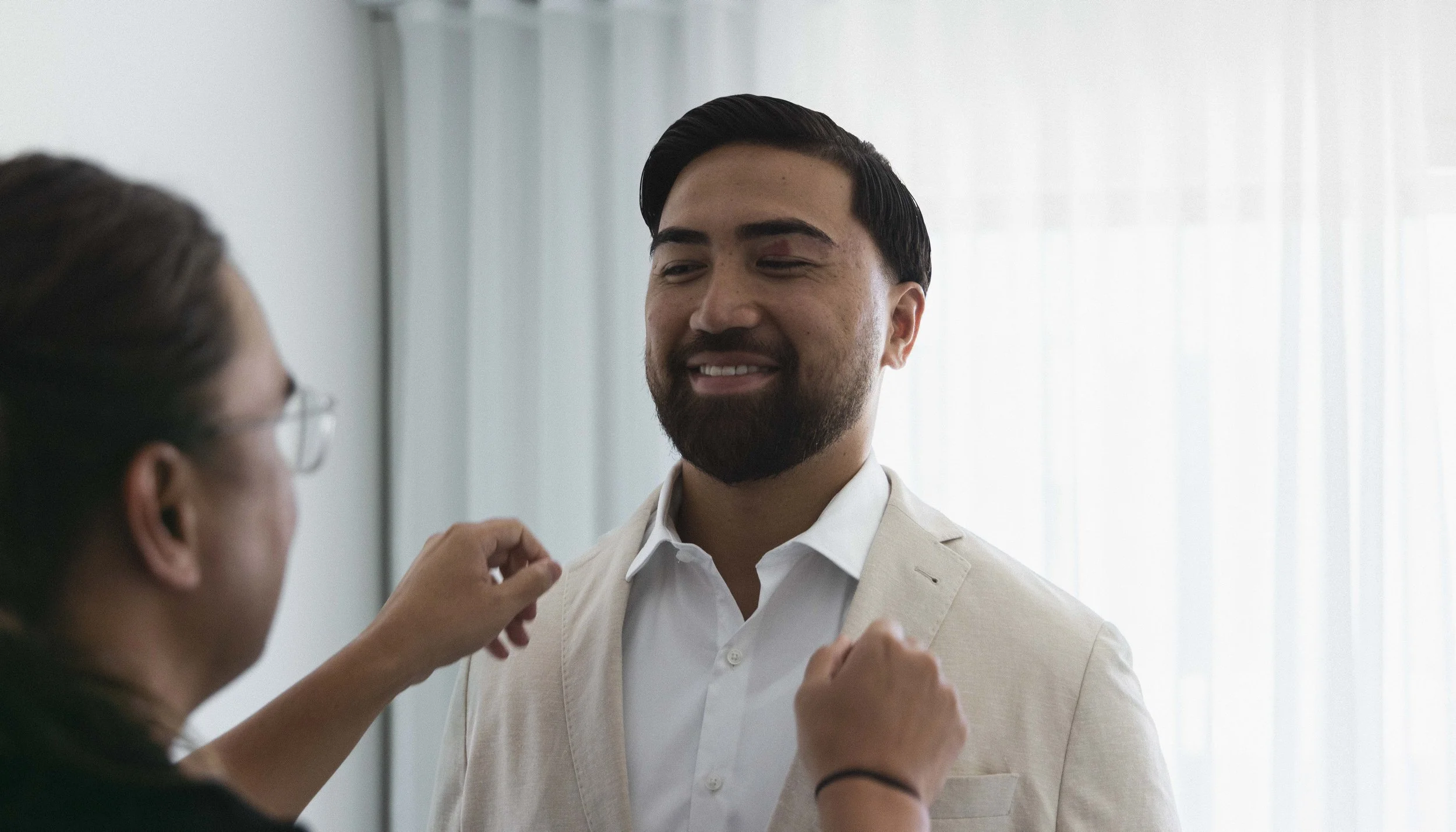 A man in a white shirt and beige blazer smiling while a woman, partially visible, adjusts his blazer.
