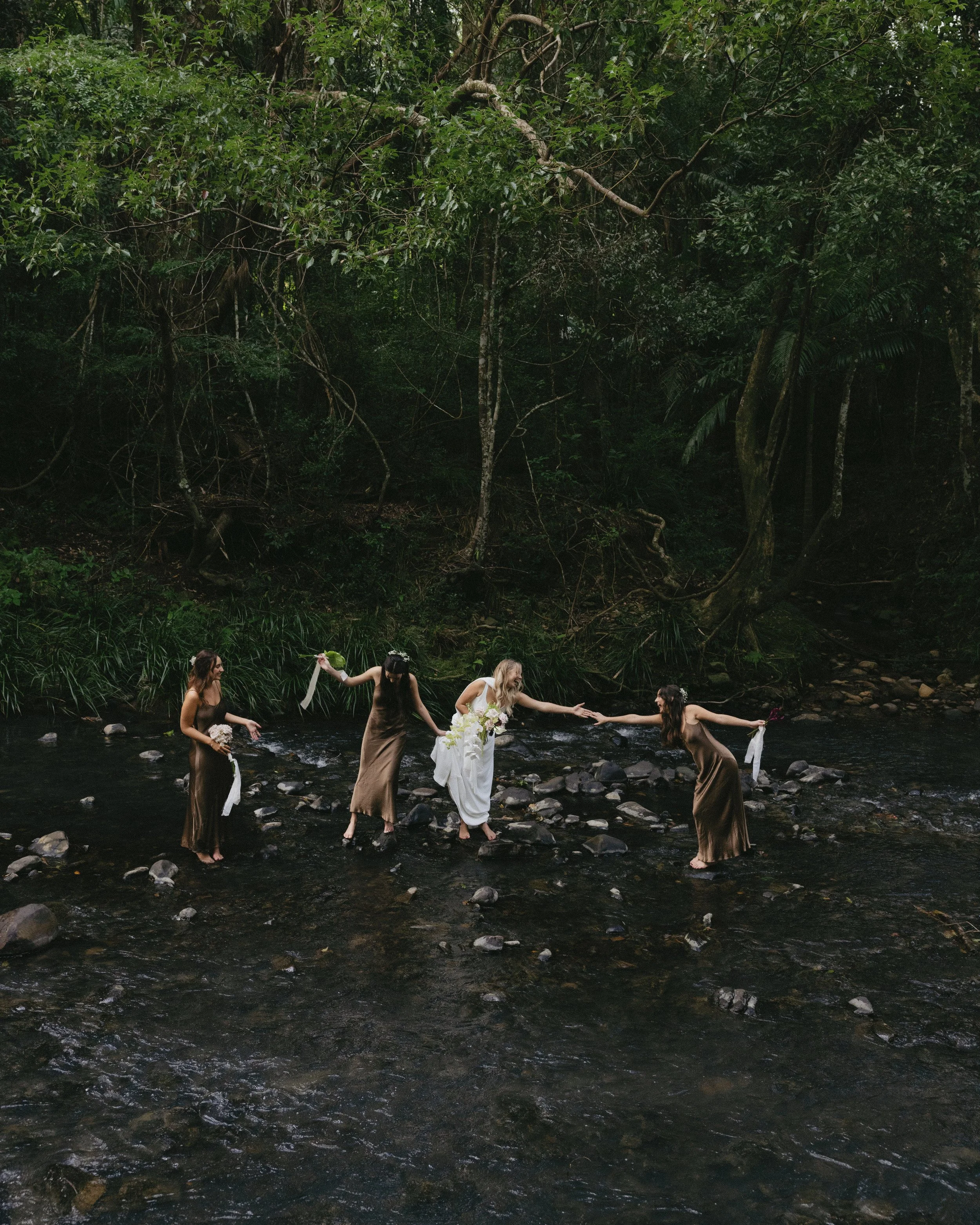 A group of five women walking on rocks in a river surrounded by dense forest, with one woman in a white dress holding flowers.