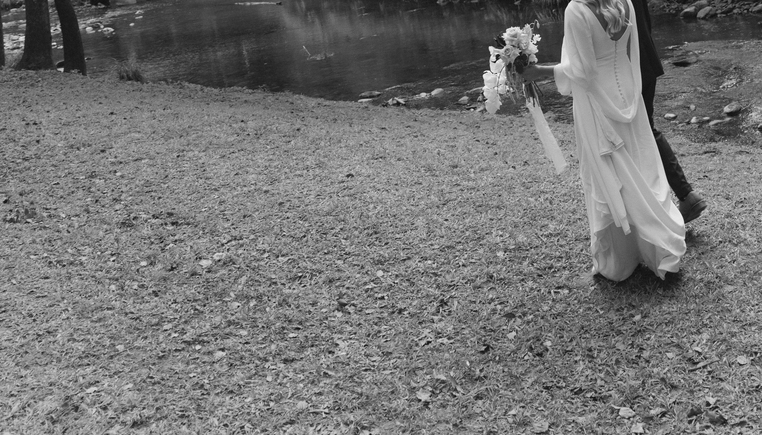 A bride holding a bouquet of flowers walking on a grassy area near a body of water, with a person in formal attire walking behind her.