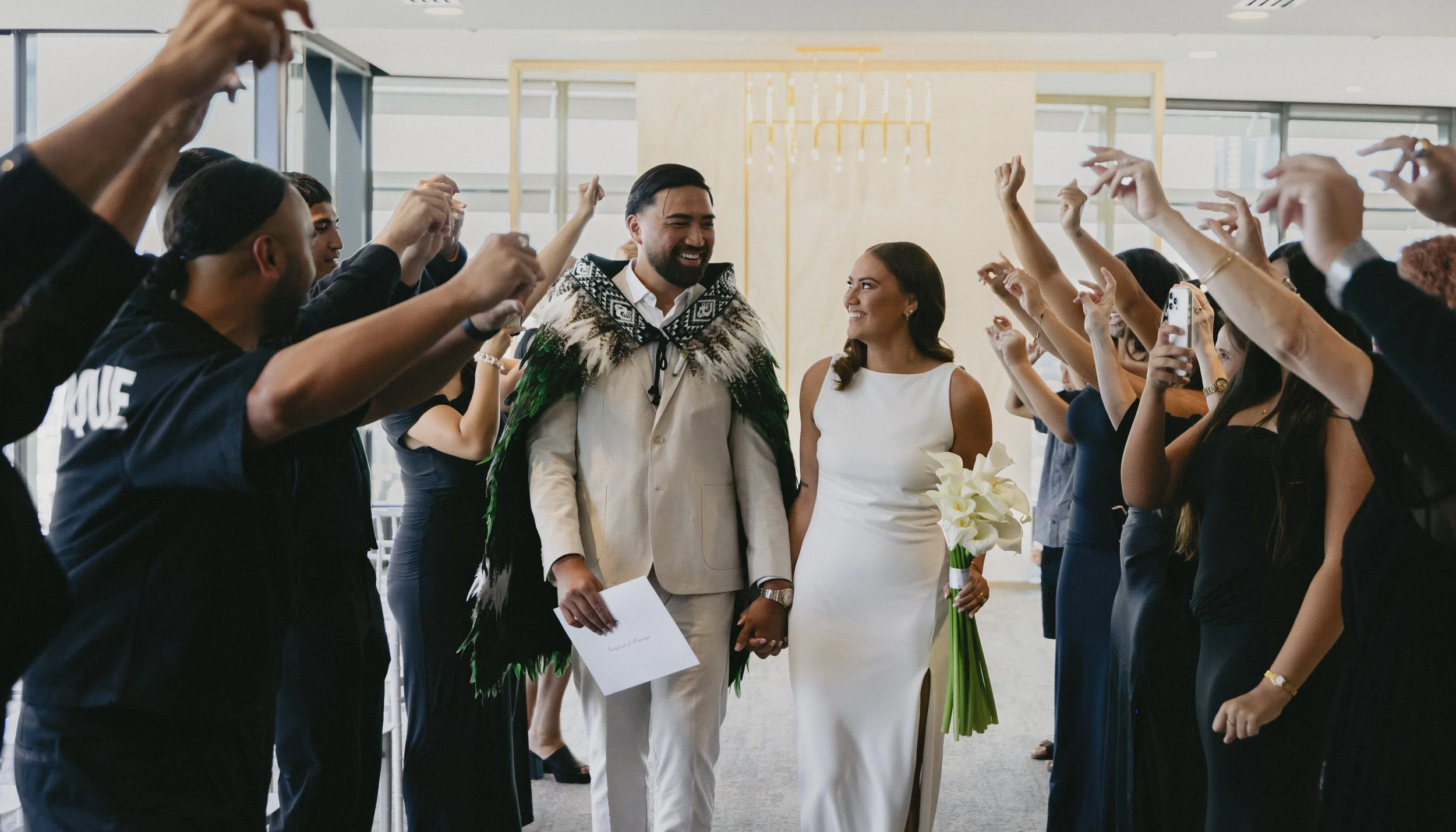 A couple dressed in wedding attire walking through a hallway on their wedding day, surrounded by friends and family raising their hands in celebration.