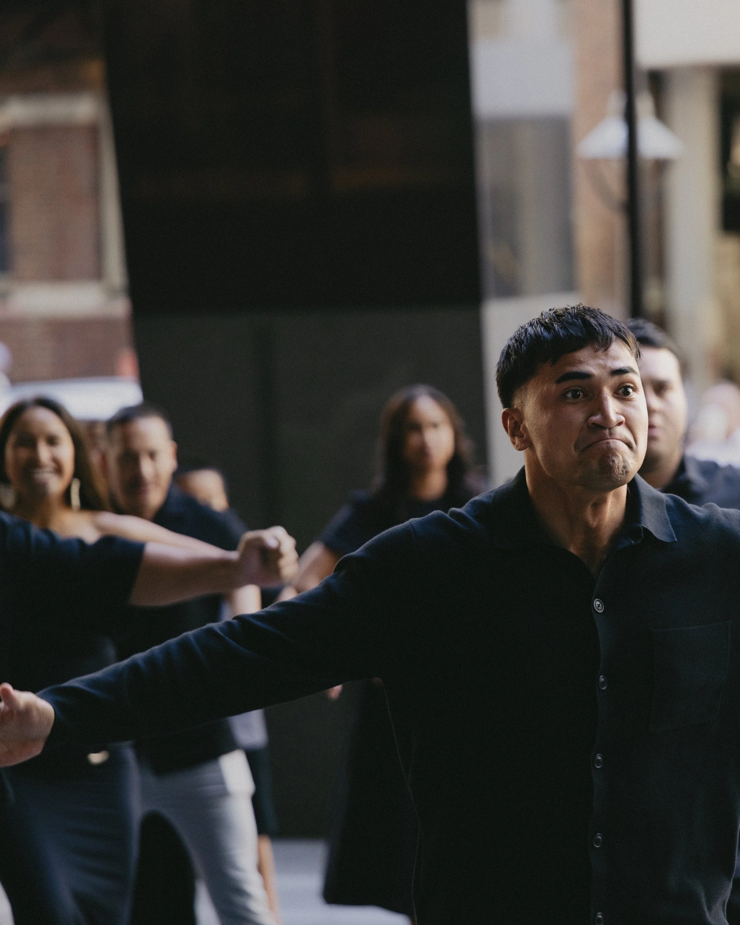 A group of people participating in a handshake or greeting, with a man in the foreground making a playful or uncertain face, outdoors in an urban setting.