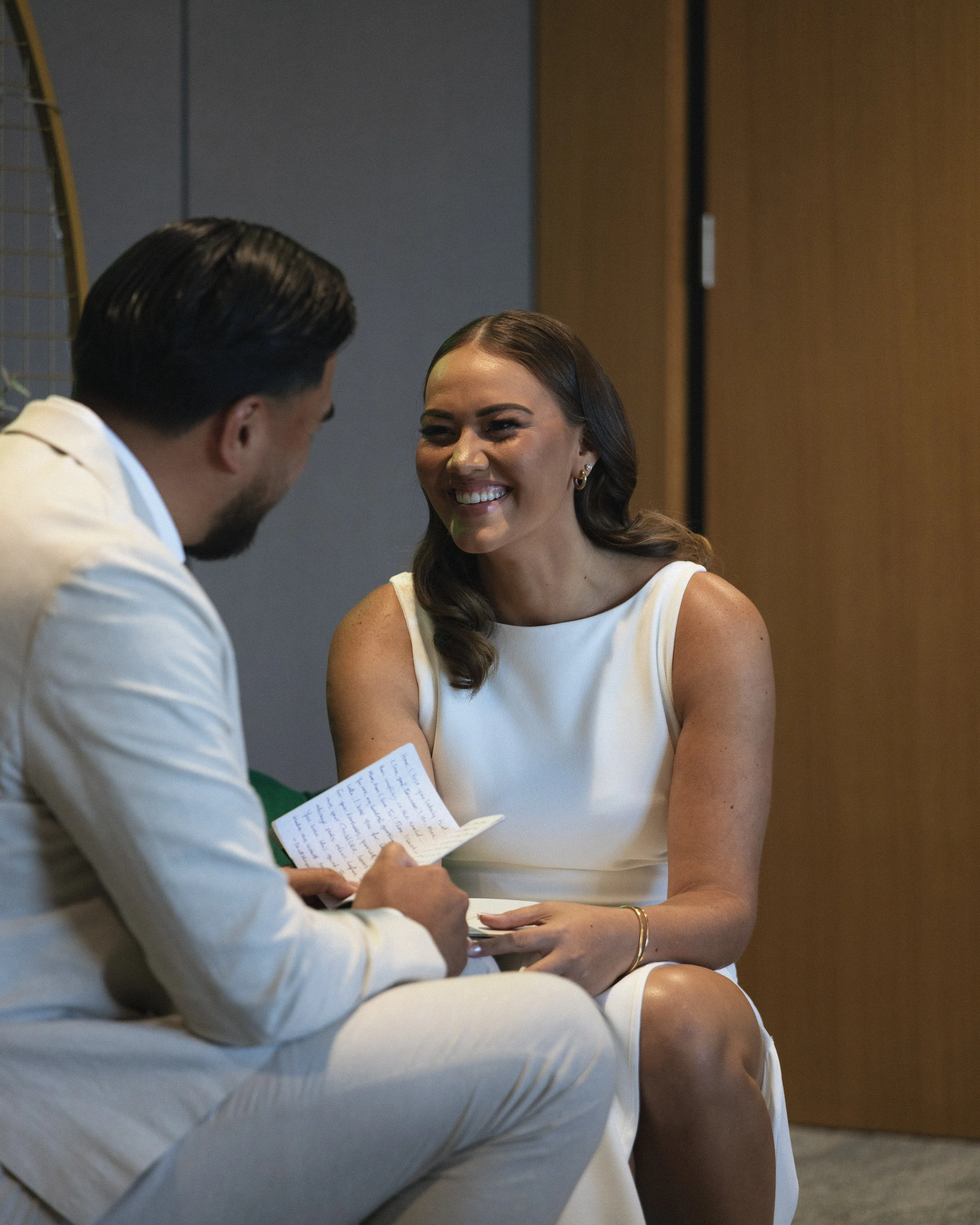 A woman in a white dress smiling and talking to a man in a white suit holding a piece of paper with text.