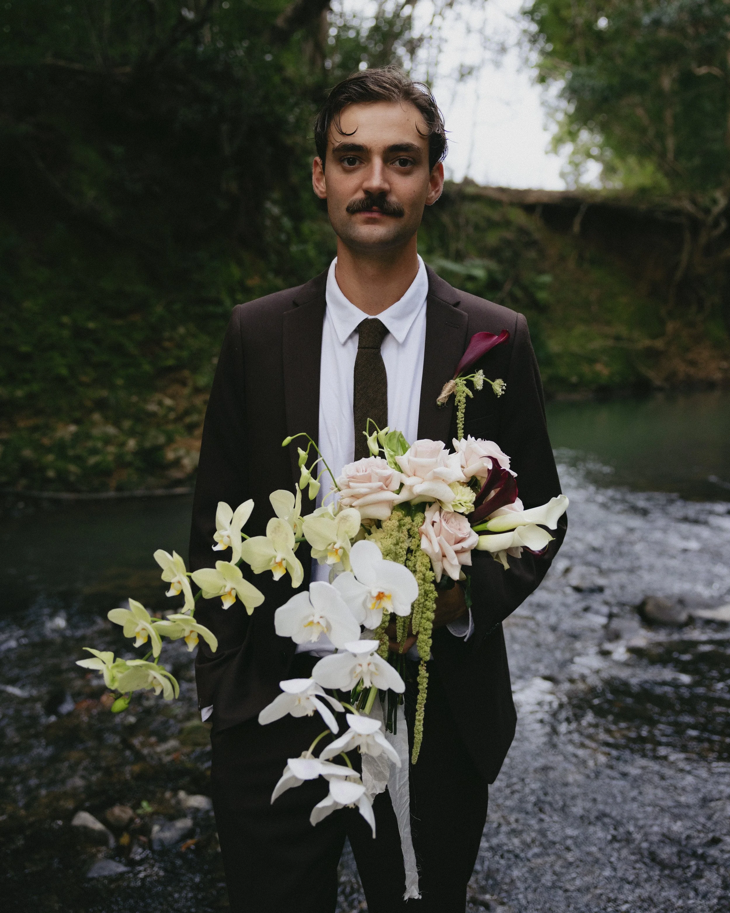 A man in a suit holding a large bouquet of assorted flowers standing by a river in a wooded area.