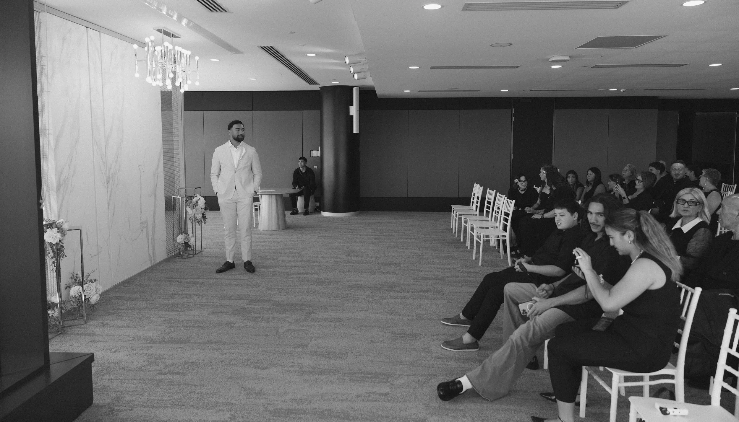 A man in a white suit stands on the left side of a room decorated with floral arrangements and a chandelier, addressing an audience seated on the right, with some taking photos or videos.