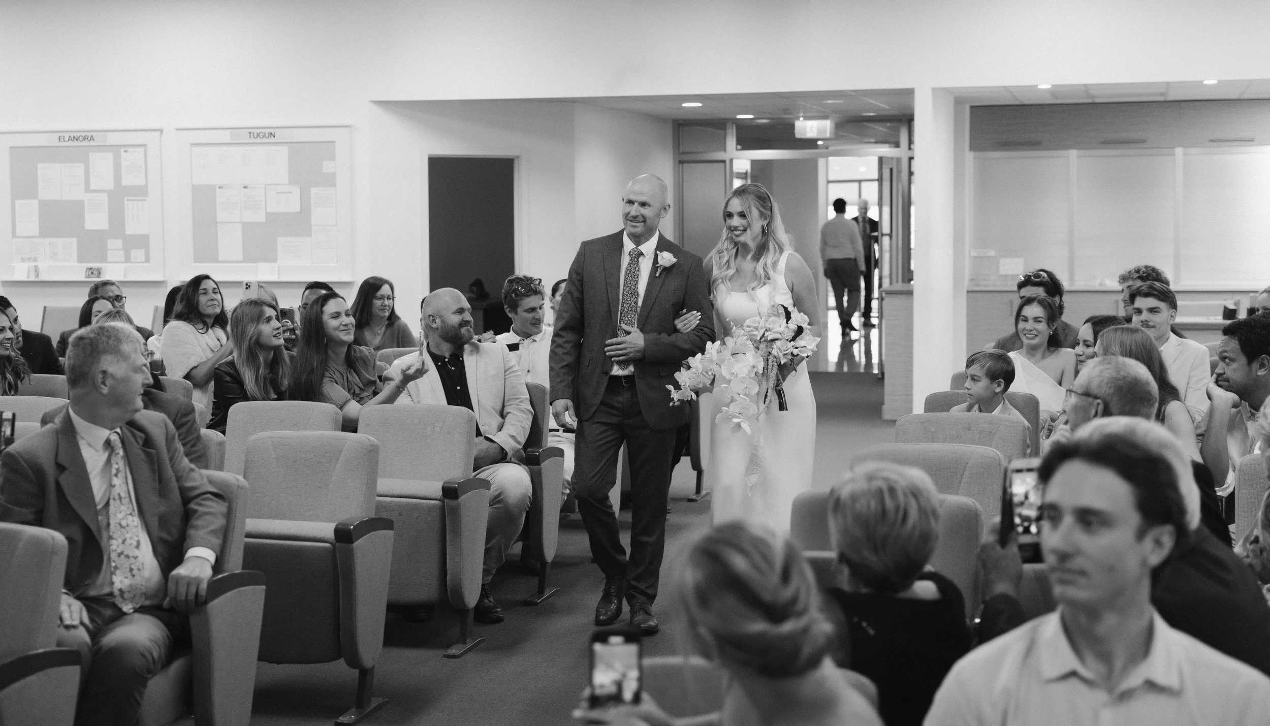 A black and white photo of a wedding ceremony. A bride in a white gown with a bouquet walks down the aisle, accompanied by a man in a suit. Guests seated on either side watch and smile, capturing photos of the moment.