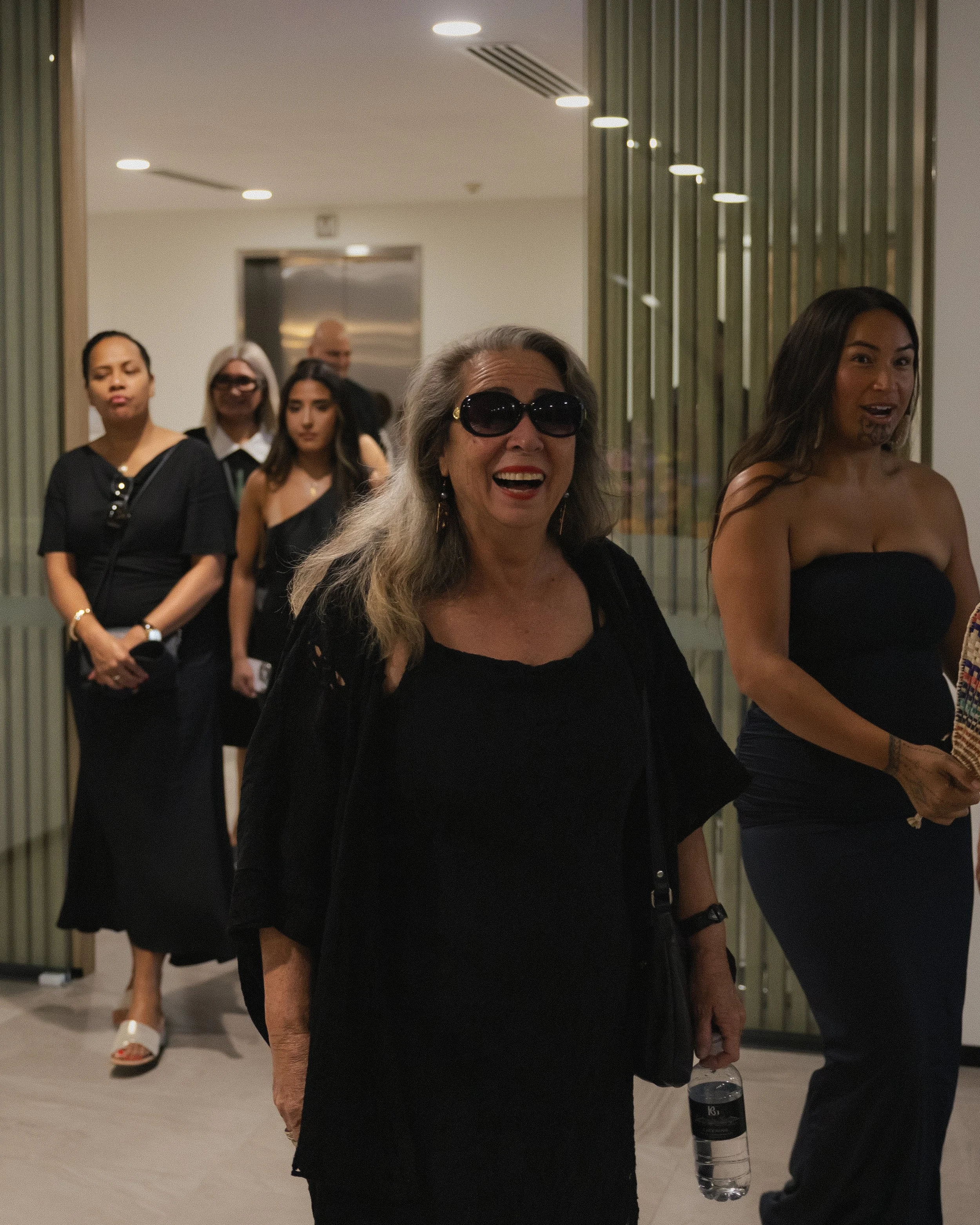 A group of women, mostly dressed in black, standing indoors with one smiling woman in the foreground wearing sunglasses and holding a water bottle.