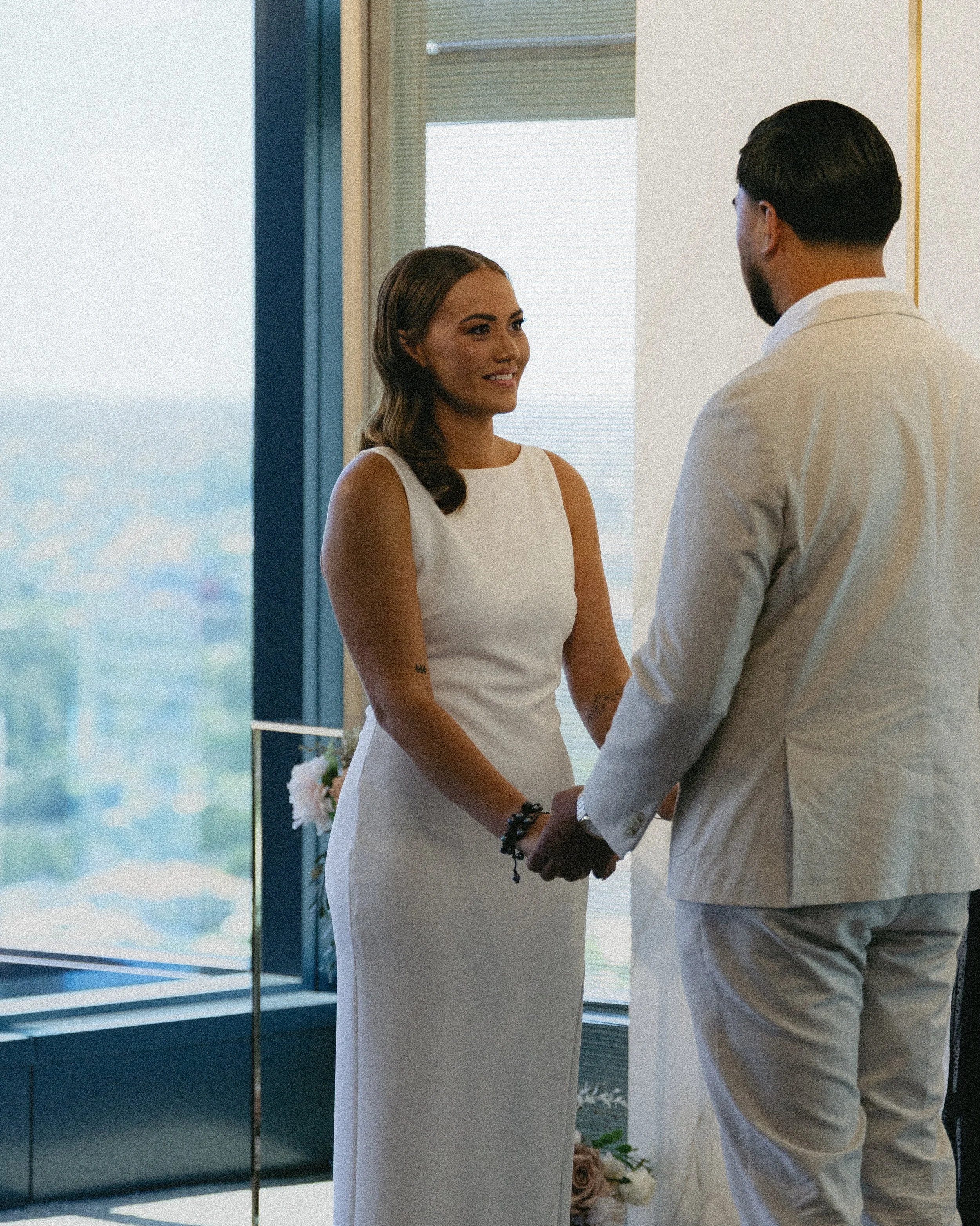 A woman and a man holding hands during a wedding ceremony indoors near a large window with city views.
