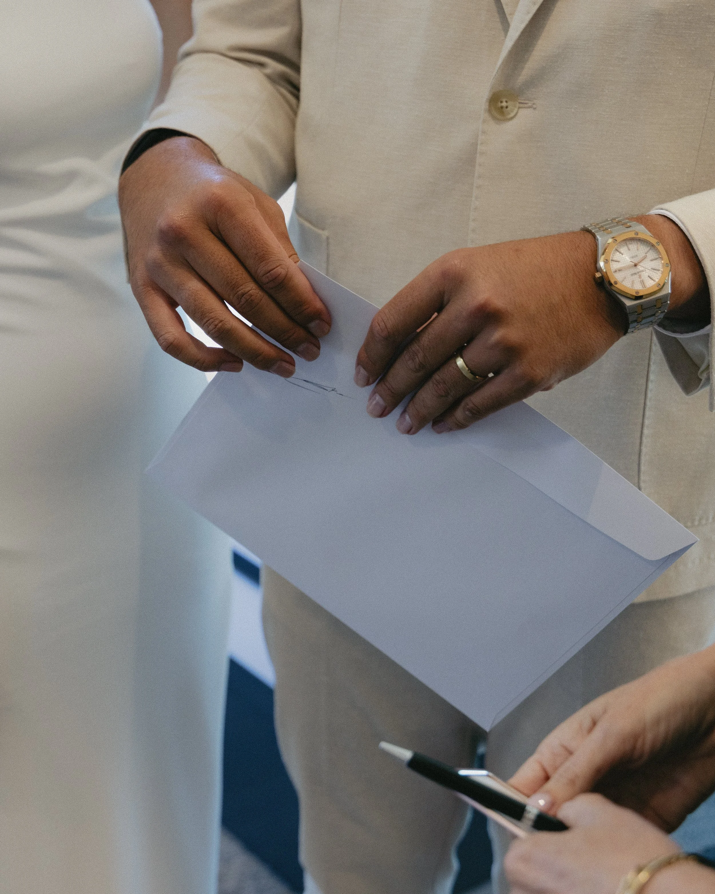 Person holding an envelope and a pen, wearing a beige suit with a silver and gold wristwatch and wedding ring.