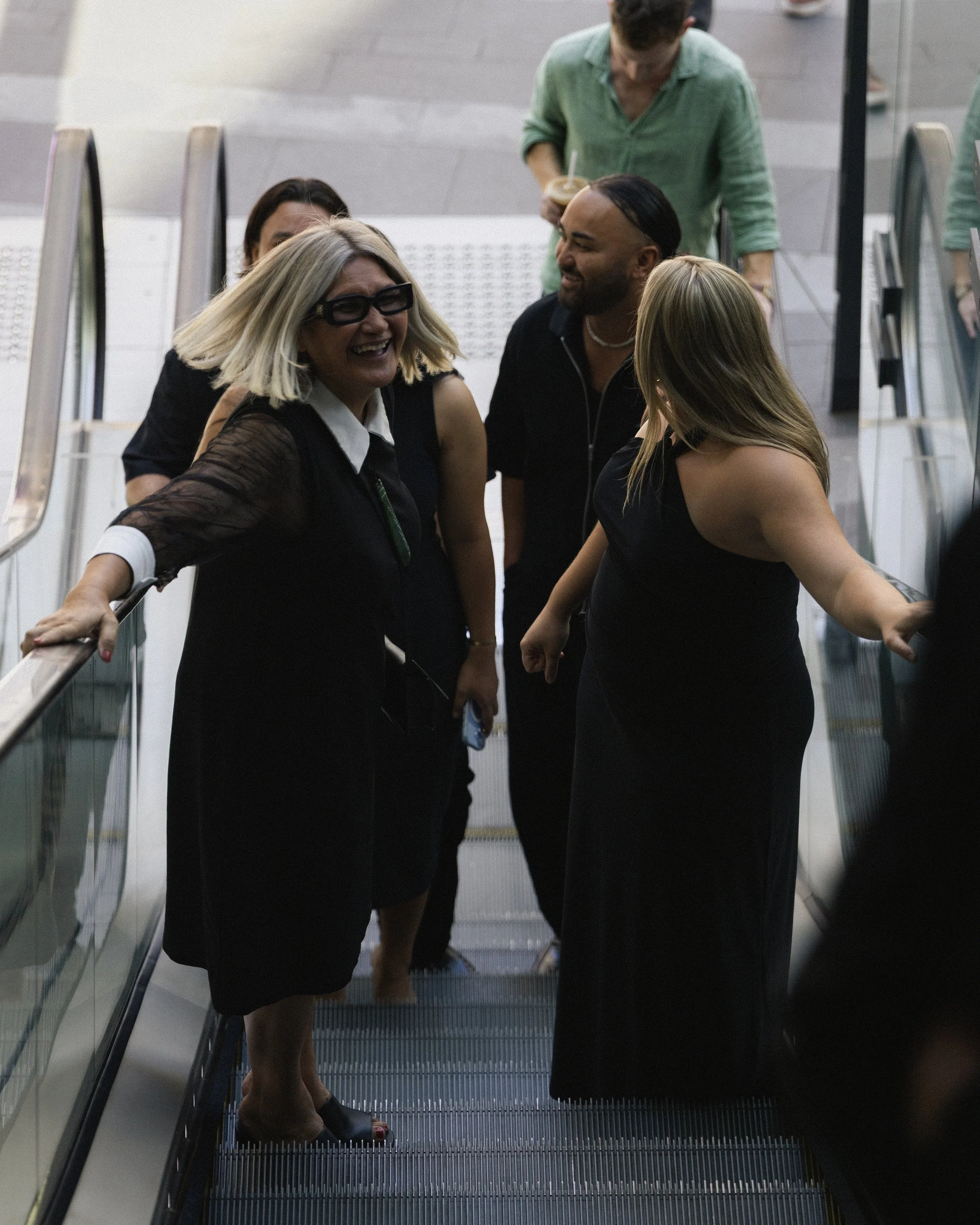 A group of people on an escalator, smiling and talking, dressed in black and casual clothing, in a public indoor space.