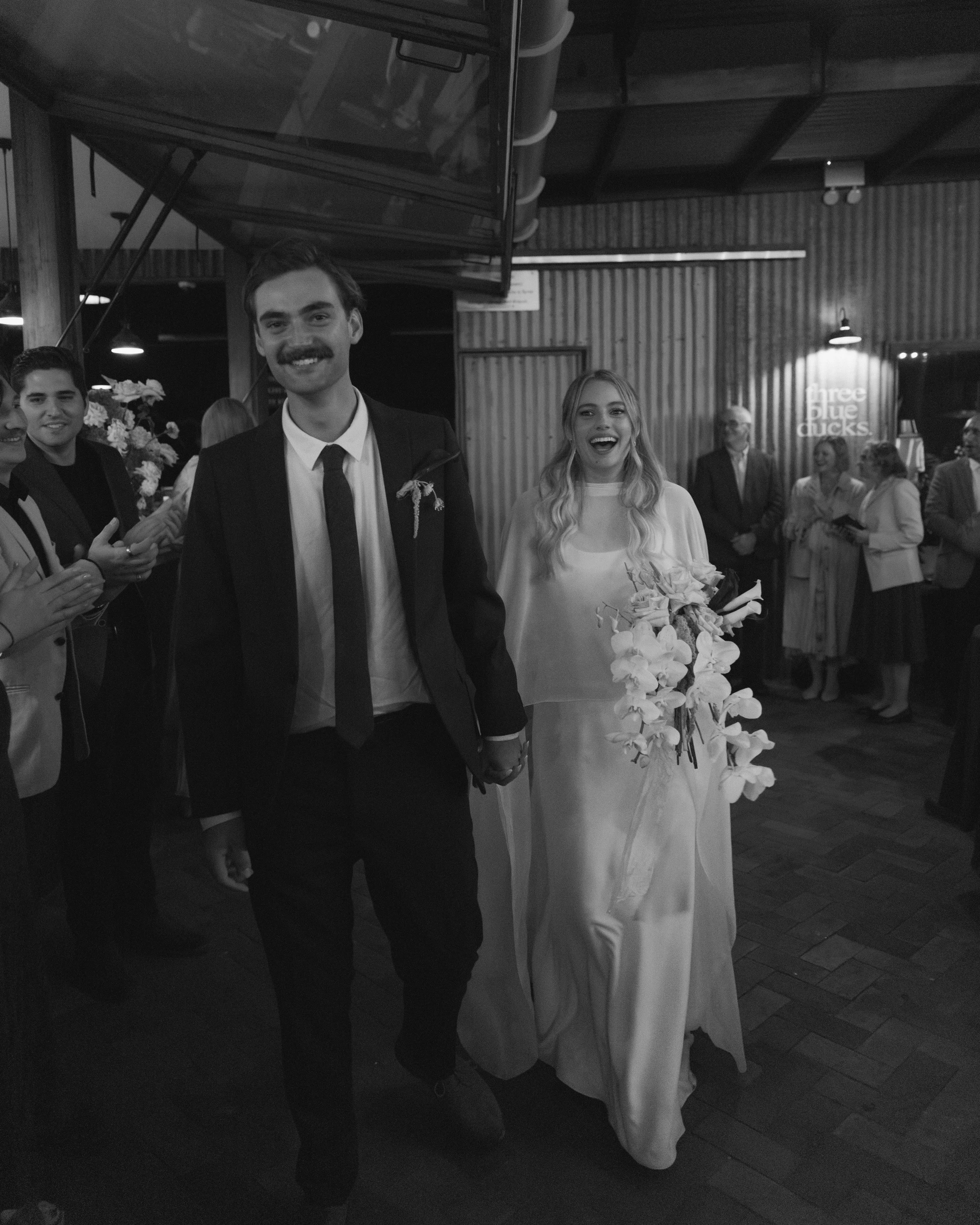 A wedding couple, the groom with a mustache in a suit and tie, and the bride holding a bouquet of flowers, walking hand-in-hand in a reception hall with guests.