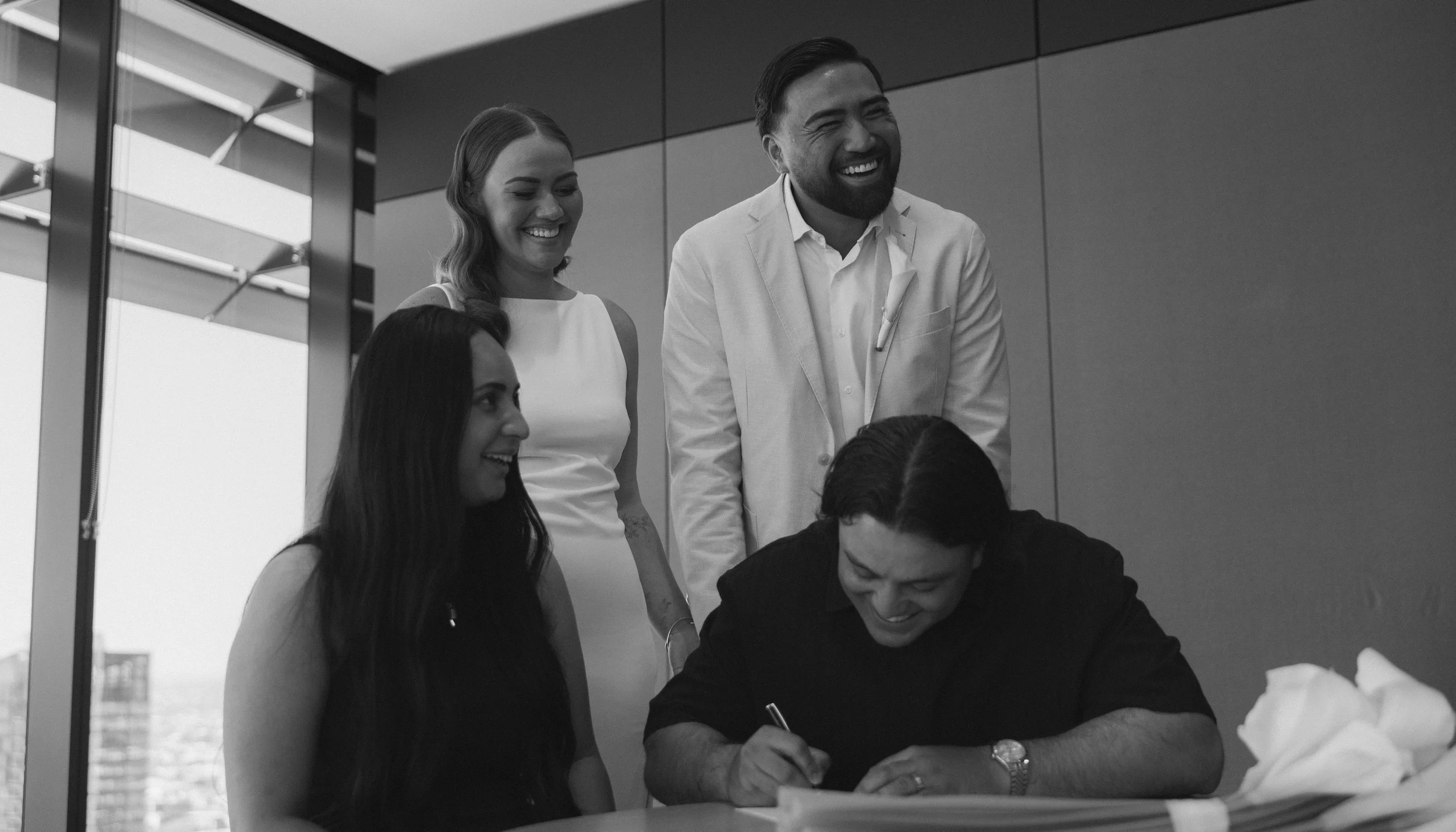 A group of four people, three women and one man, are gathered around a table watching as a man signs a document. They are all smiling and appear to be celebrating or enjoying the moment in a modern office.