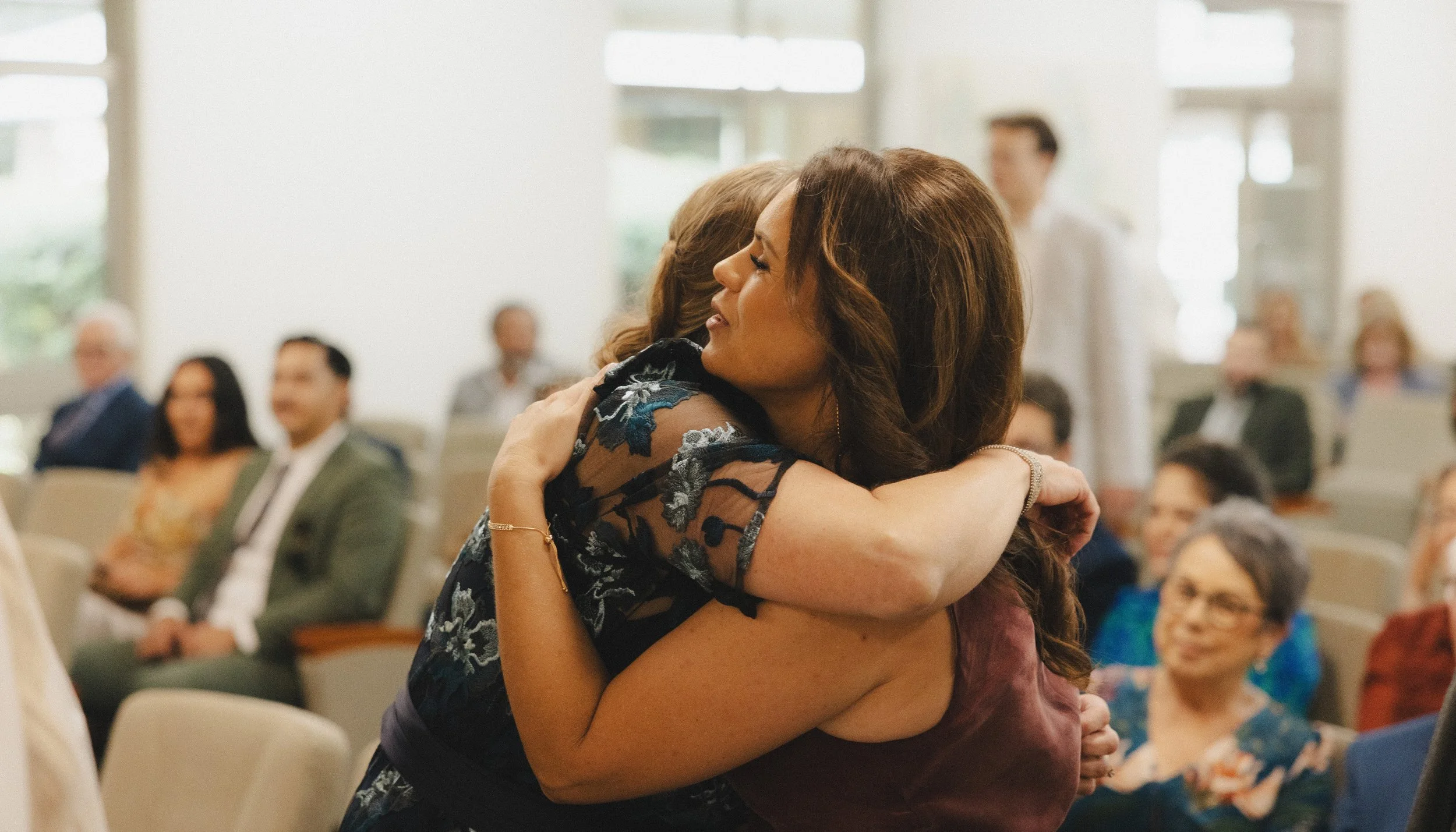 Two women hugging in a gathering or ceremony, with several seated and standing people in the background.