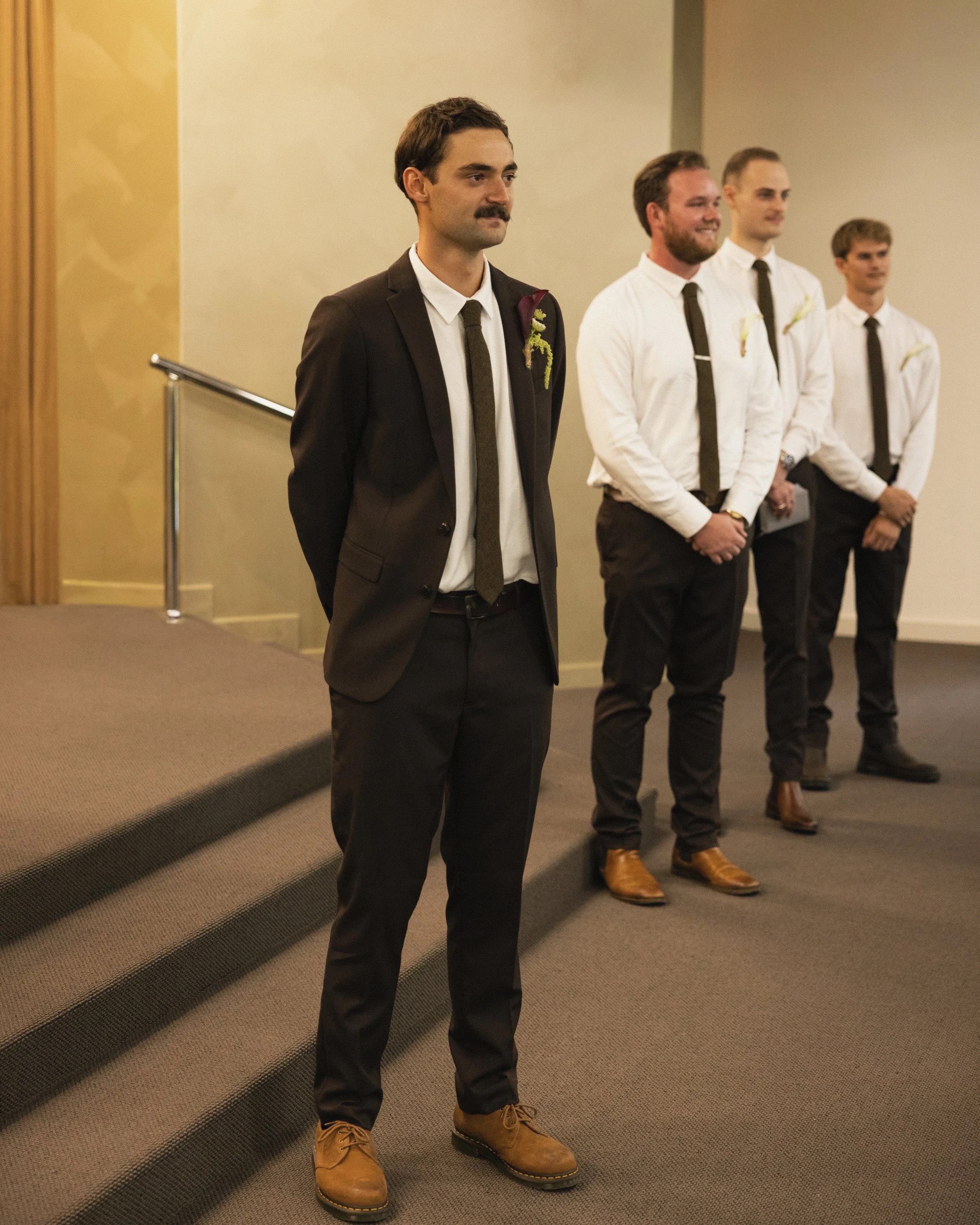 Four men in formal attire standing in a line with hands clasped, attending a formal event or ceremony in a room with beige walls and carpeted floor.