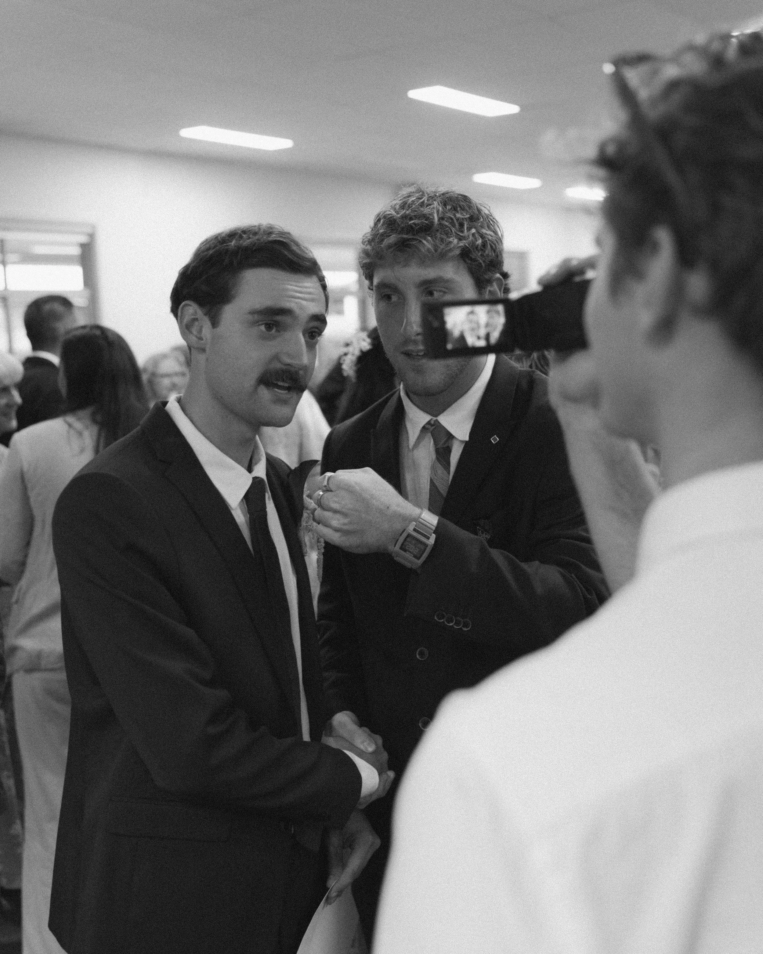 Three men in suits and ties are talking and laughing at a social event, with a woman nearby taking a photo of them, in a room filled with other people.
