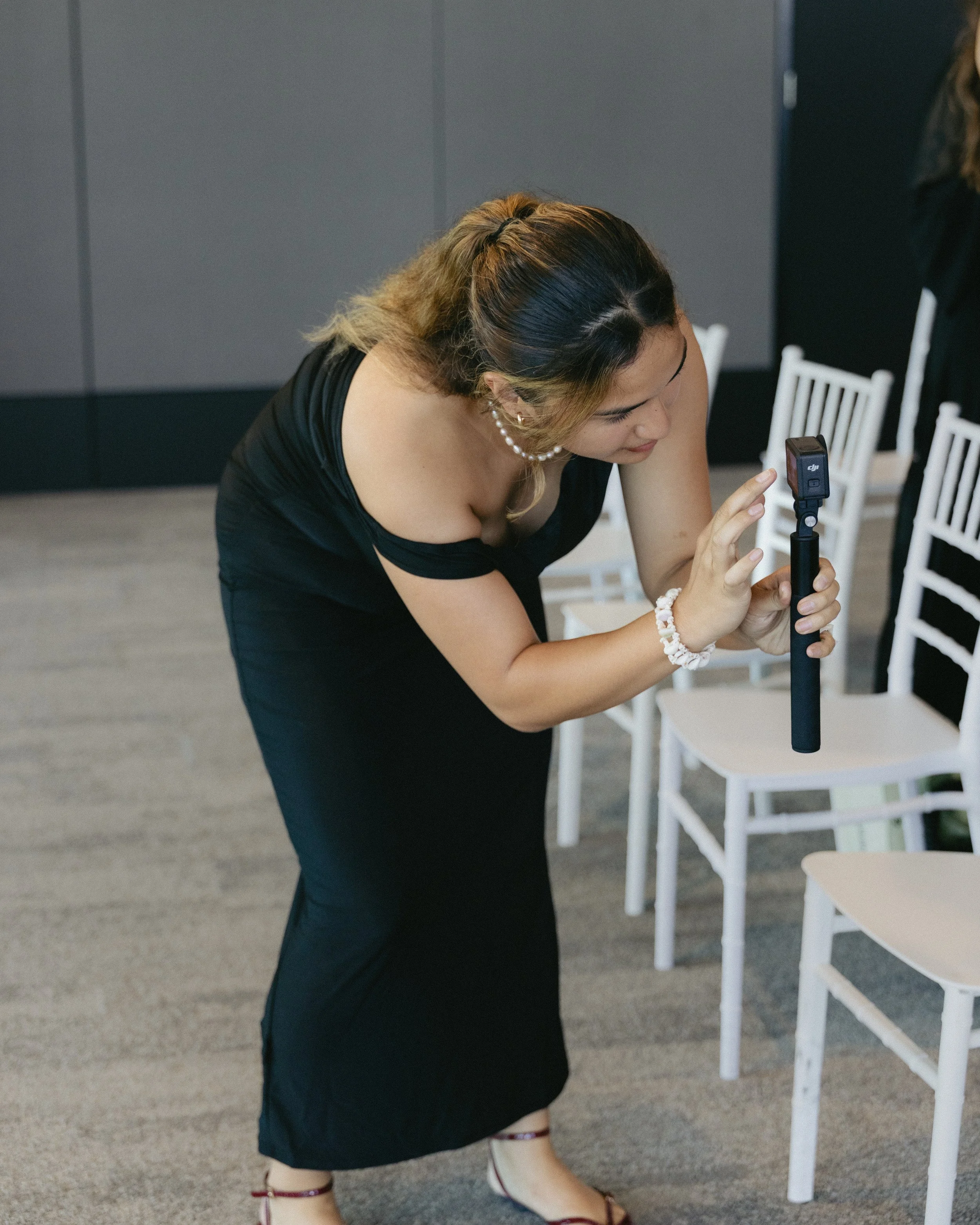 Woman in black dress taking a photo with a selfie stick in a room with white chairs.