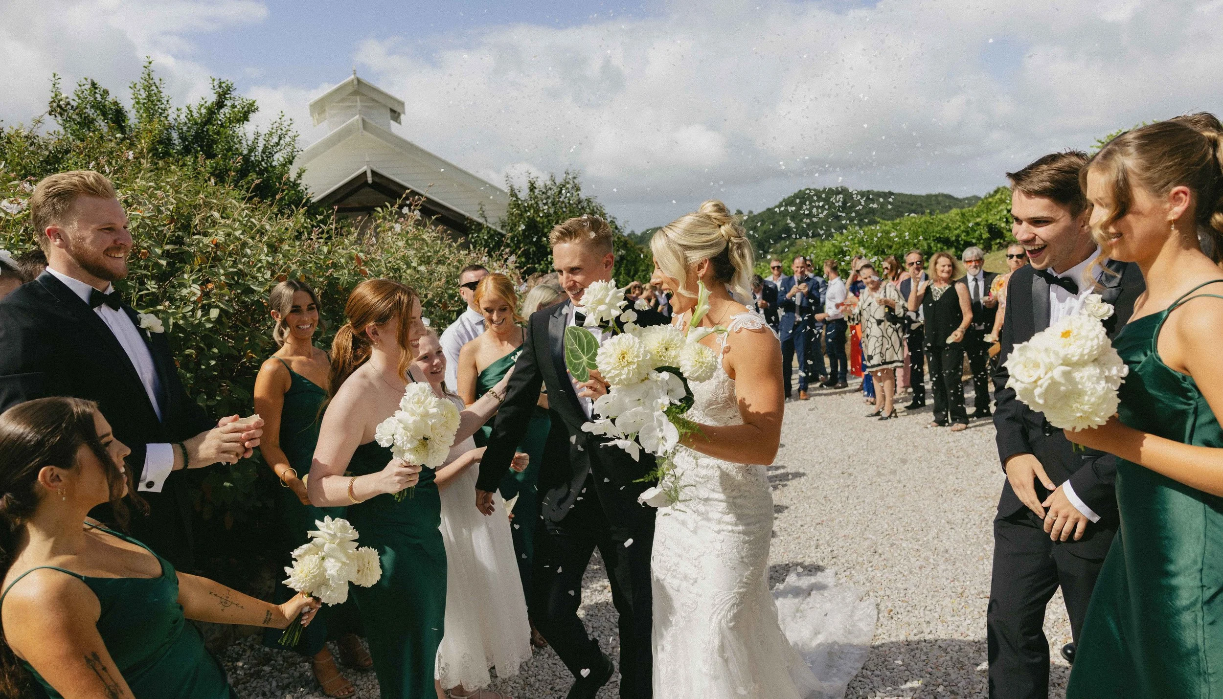 A wedding celebration outdoors with a bride and groom walking through a line of guests throwing flower petals and confetti. The bride is wearing a white wedding dress and carrying a bouquet of white flowers, while the groom is dressed in a black tuxedo. The guests, dressed in formal attire, are smiling and celebrating.