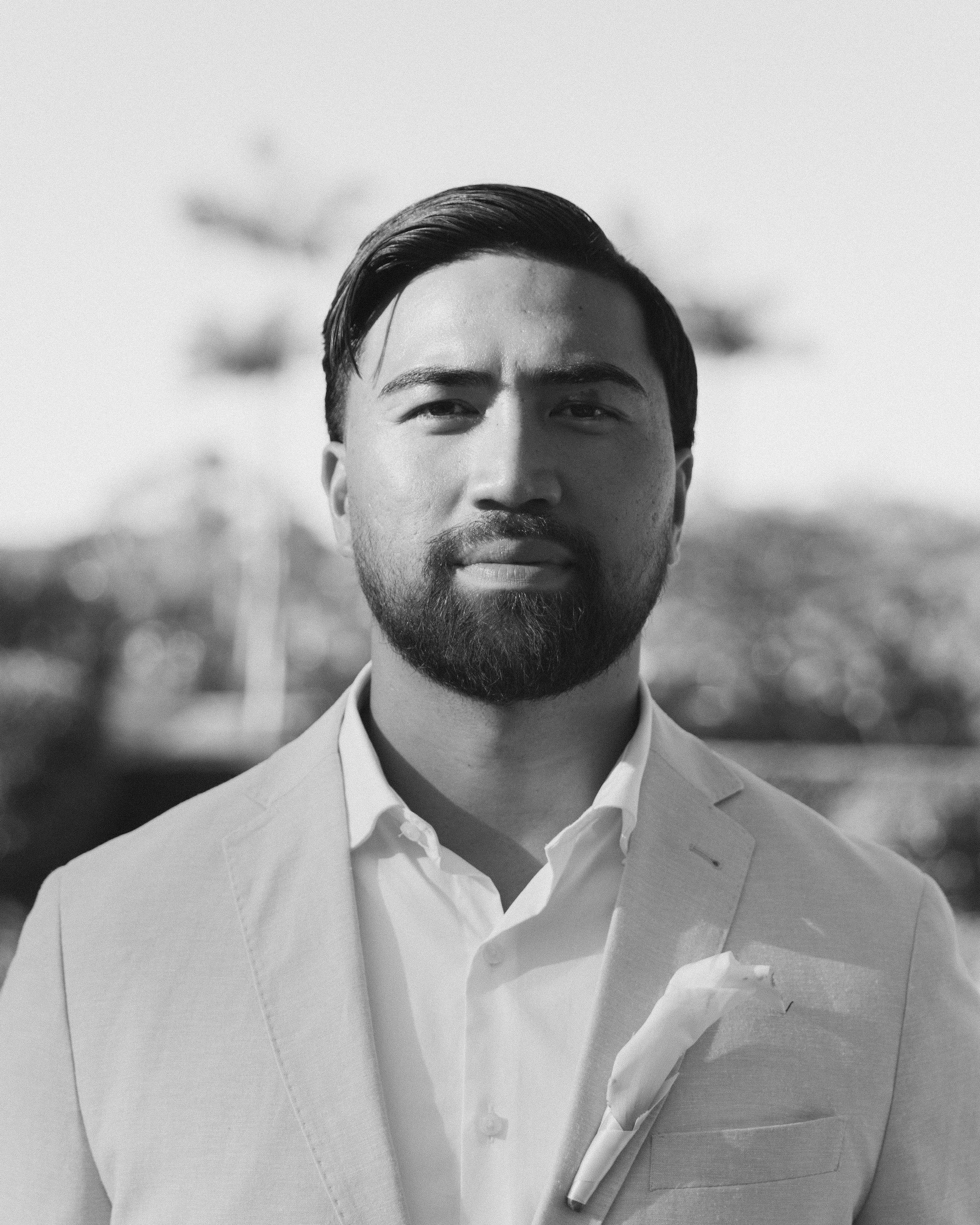 Black and white portrait of a man with neatly combed hair, beard, and mustache, wearing a light-colored suit and shirt, outdoors with blurred background and birds in flight.