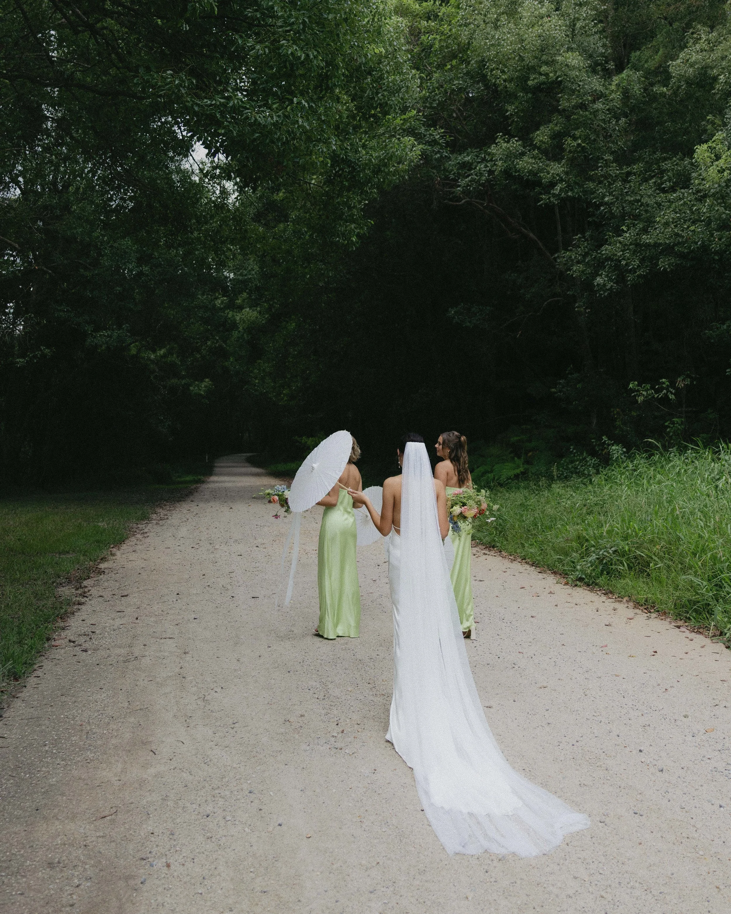 A bride with a long white train and veil walking on a dirt path in a wooded area, accompanied by two bridesmaids holding bouquets and one holding an umbrella, all dressed in light green dresses.