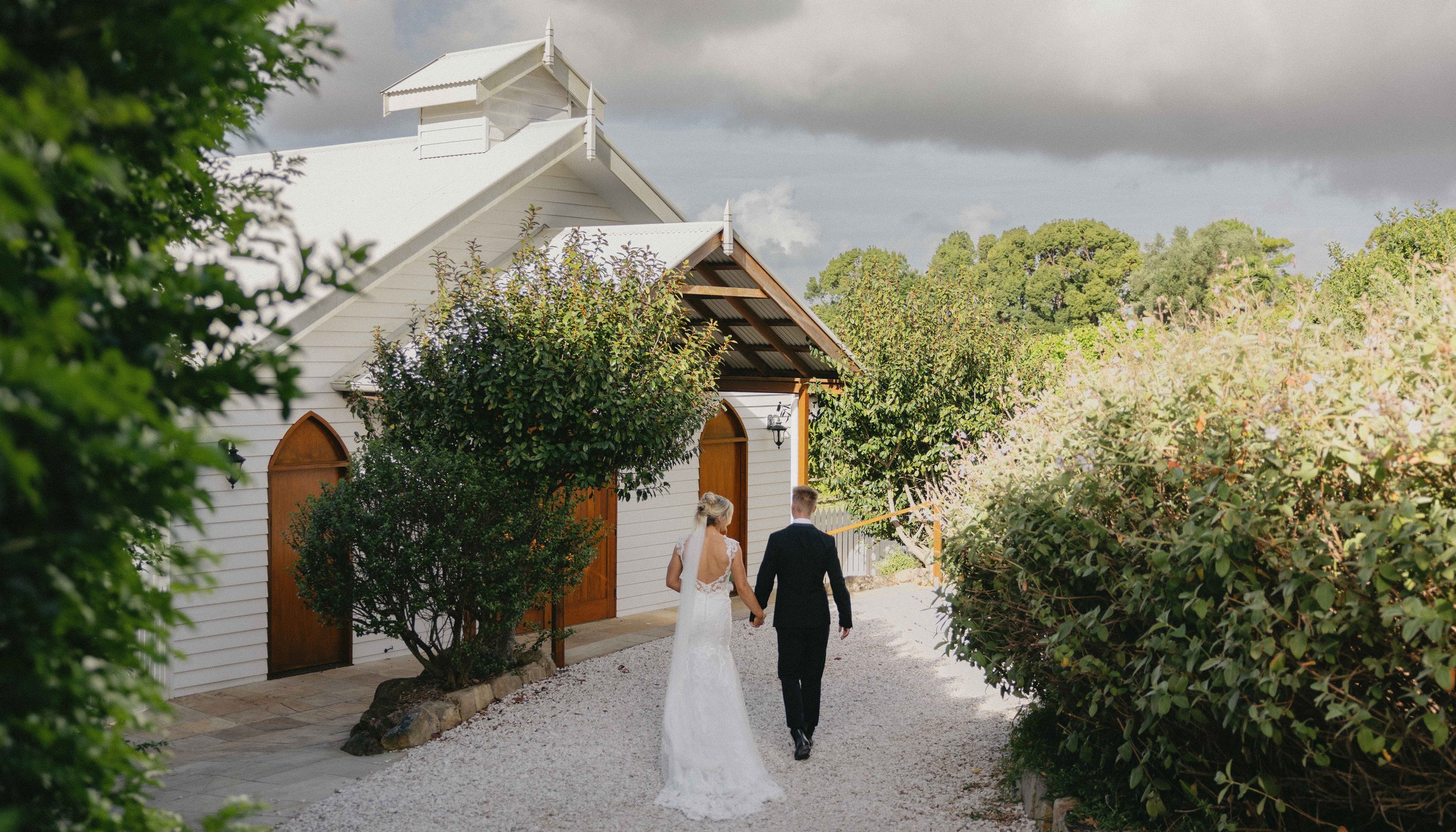 A bride and groom walking hand in hand outside a white wooden church with brown doors, surrounded by trees and bushes, on a cloudy day.