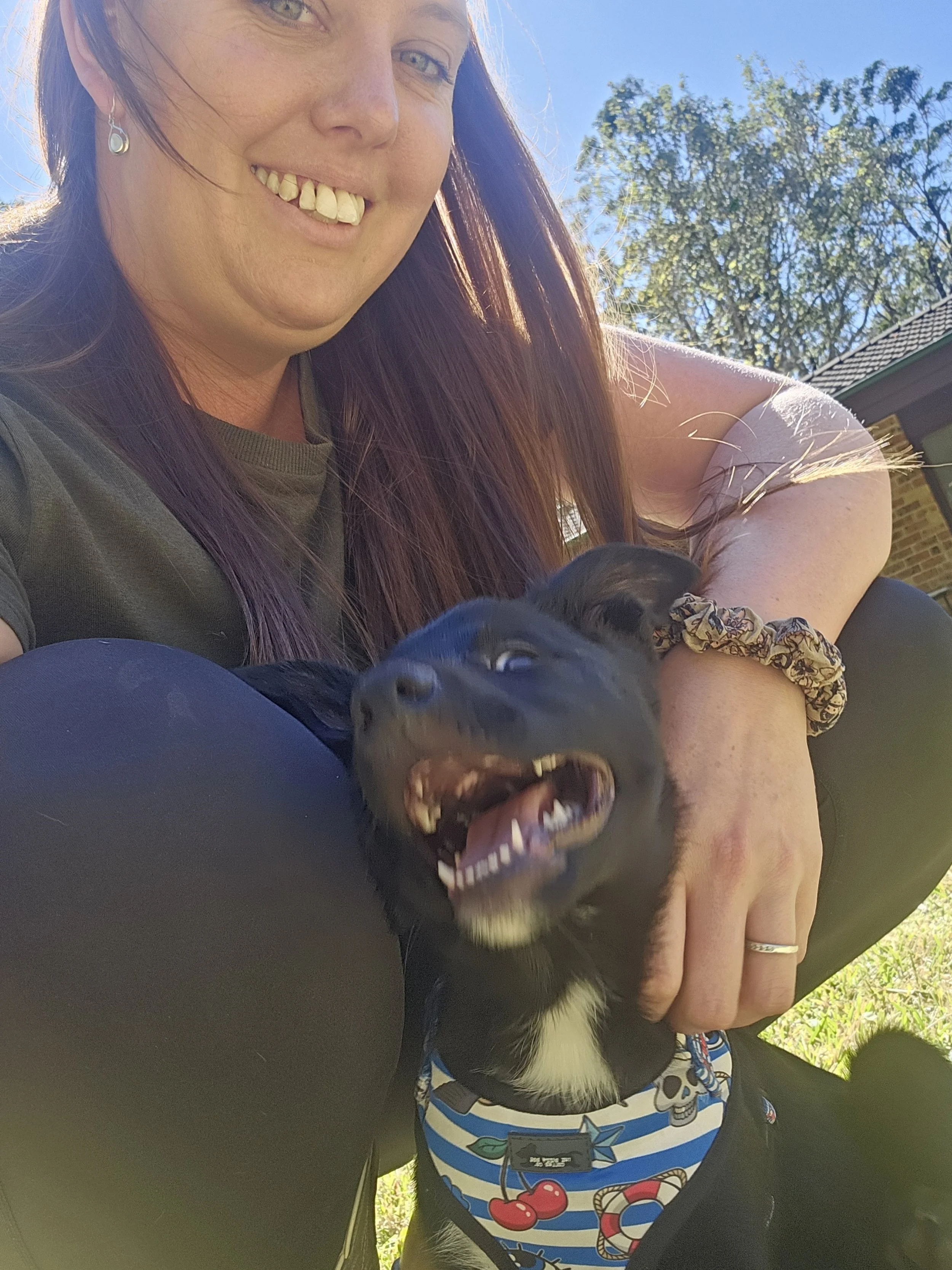 A woman with long red hair smiling outdoors while holding a black puppy with a striped blue, white, and red bandana around its neck. The puppy appears to be playfully barking or biting, with its mouth open showing teeth. The woman is wearing a ring on her finger and an earring, and the background includes green trees and a house with a brick wall.