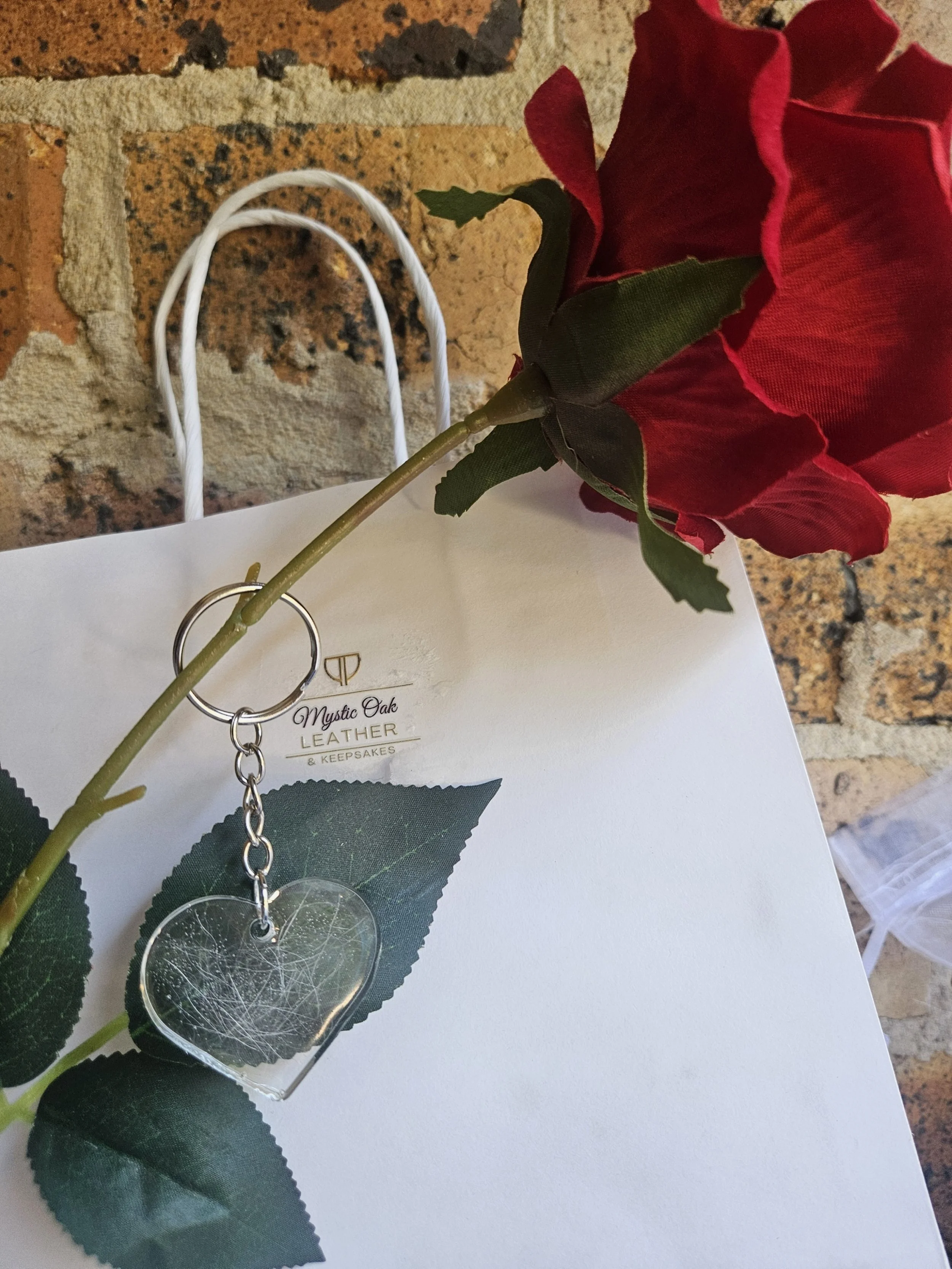 A red artificial rose with green leaves lying over a white card, with a clear heart-shaped keychain showing white fur encased and a single chain link attached, on a brick wall background.