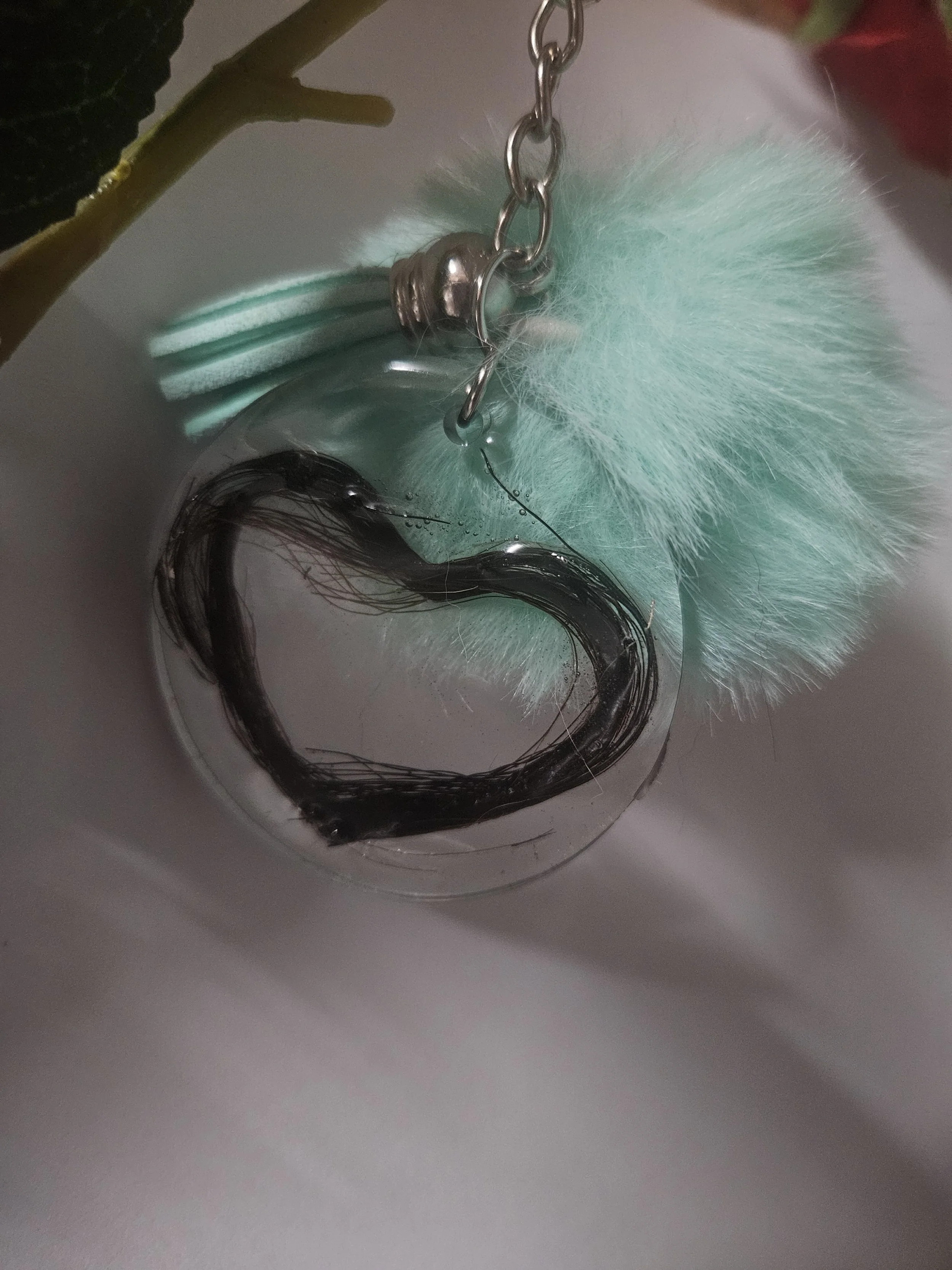 A close-up of a keychain with a black hair ring and a fluffy light blue pom-pom attached to a silver chain.
