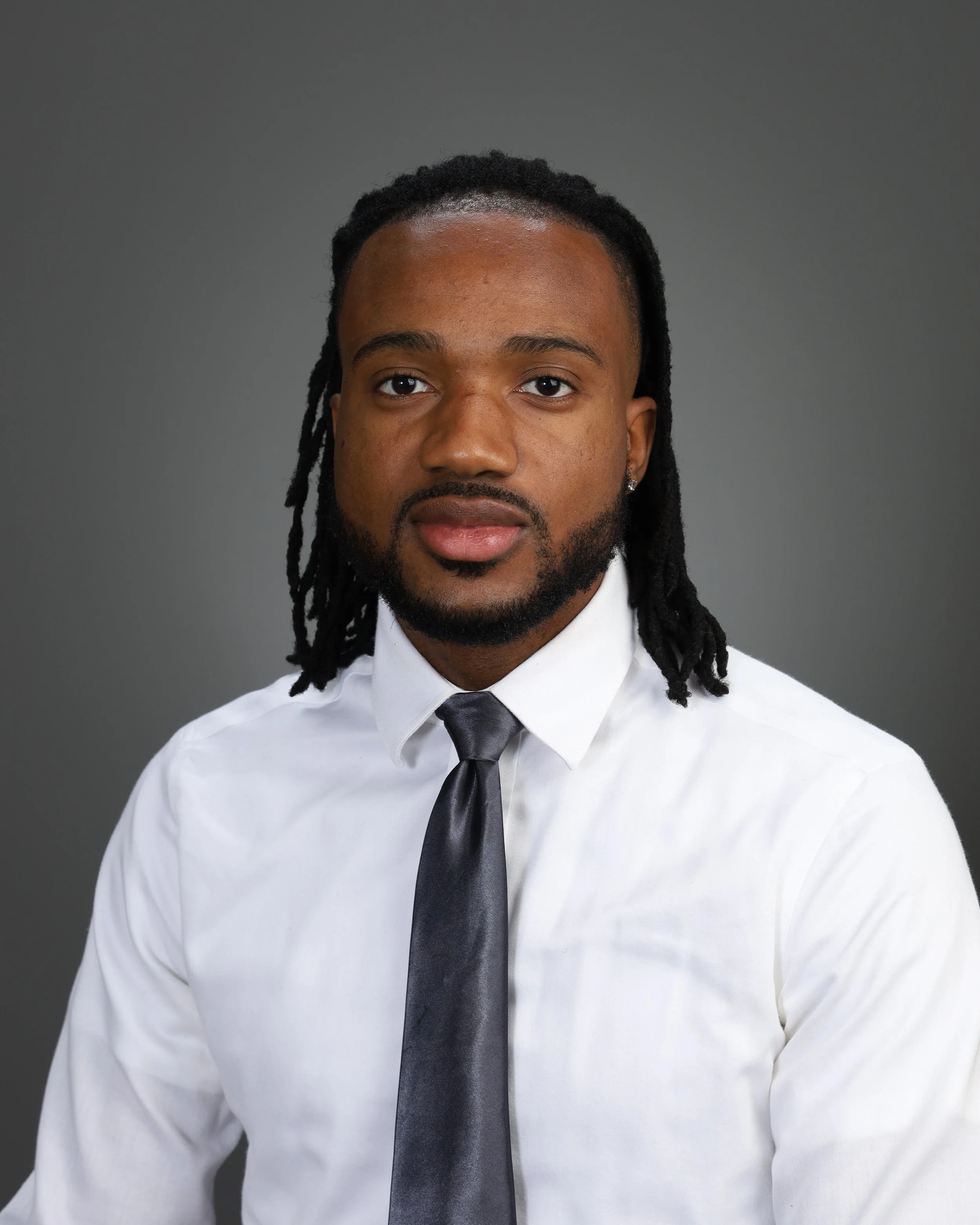 Professional headshot of a man with dreadlocks, wearing a white shirt and a dark tie, against a gray background.