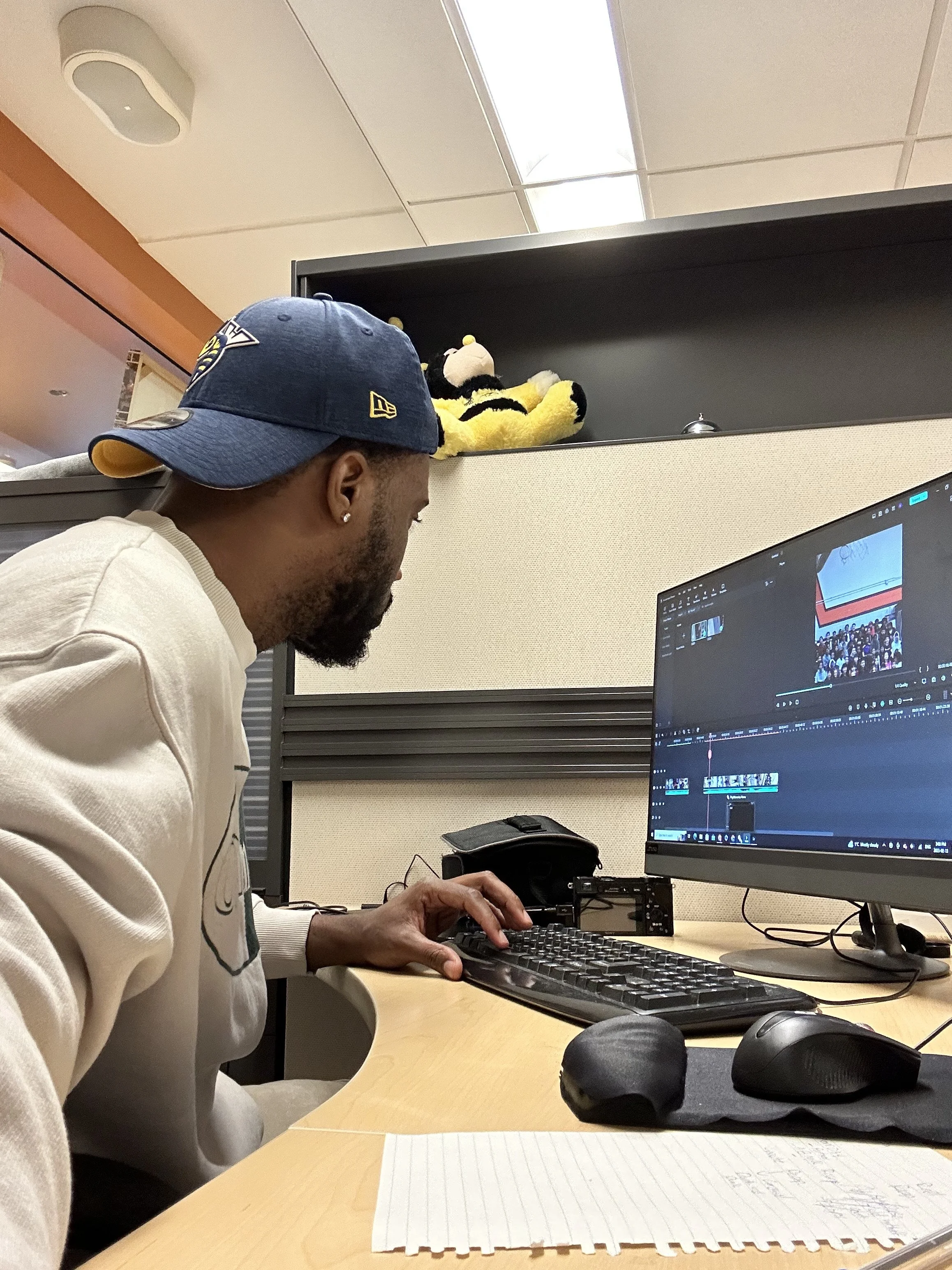 A person editing a video on a computer in an office cubicle with a computer monitor, keyboard, mouse, and paper with handwritten notes, and stuffed animal toys on the cubicle wall.