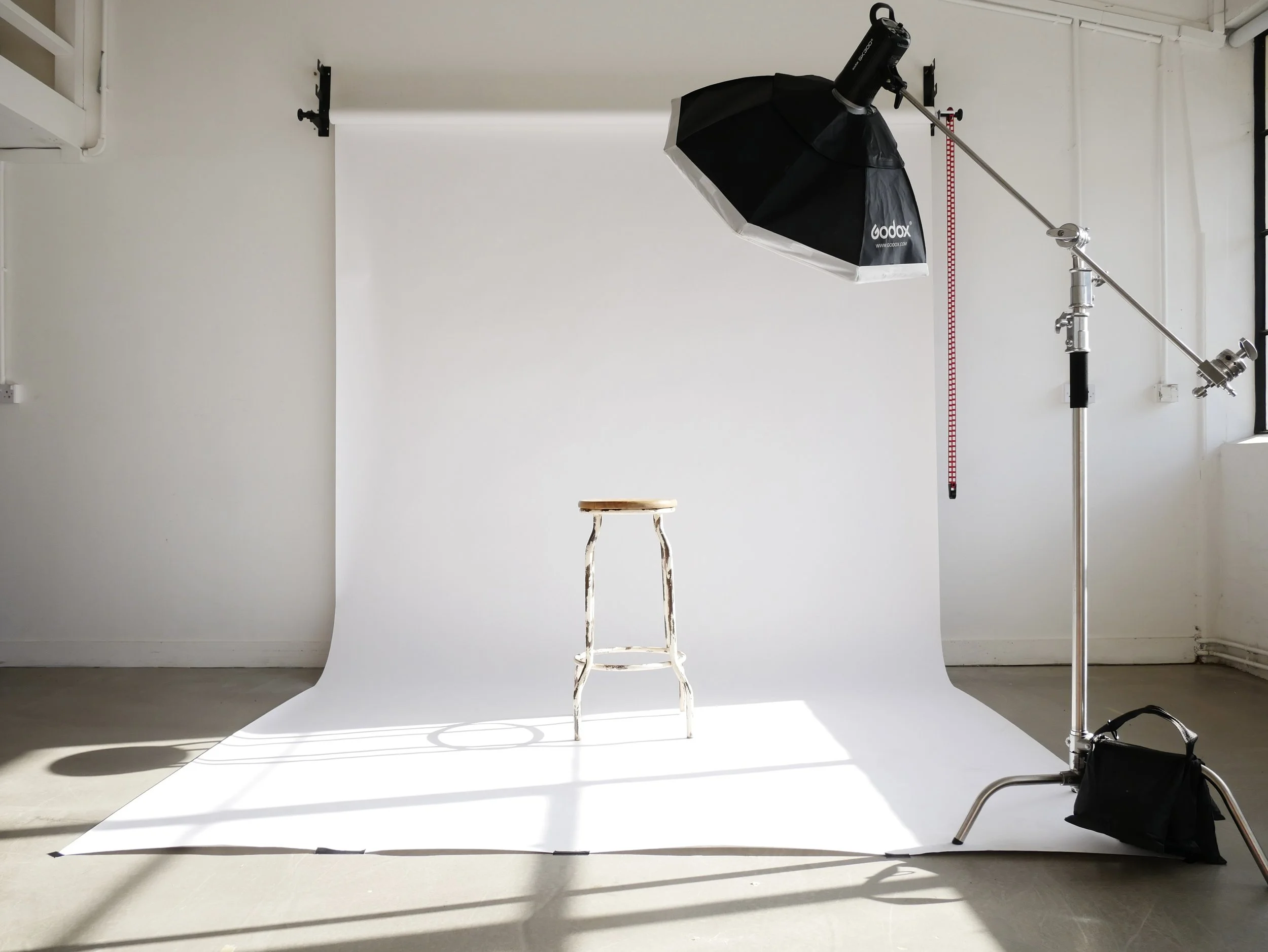 Photography studio setup with a white seamless backdrop, a wooden stool in front, large studio light with umbrella modifier on the right, and a red measuring tape hanging on the wall.