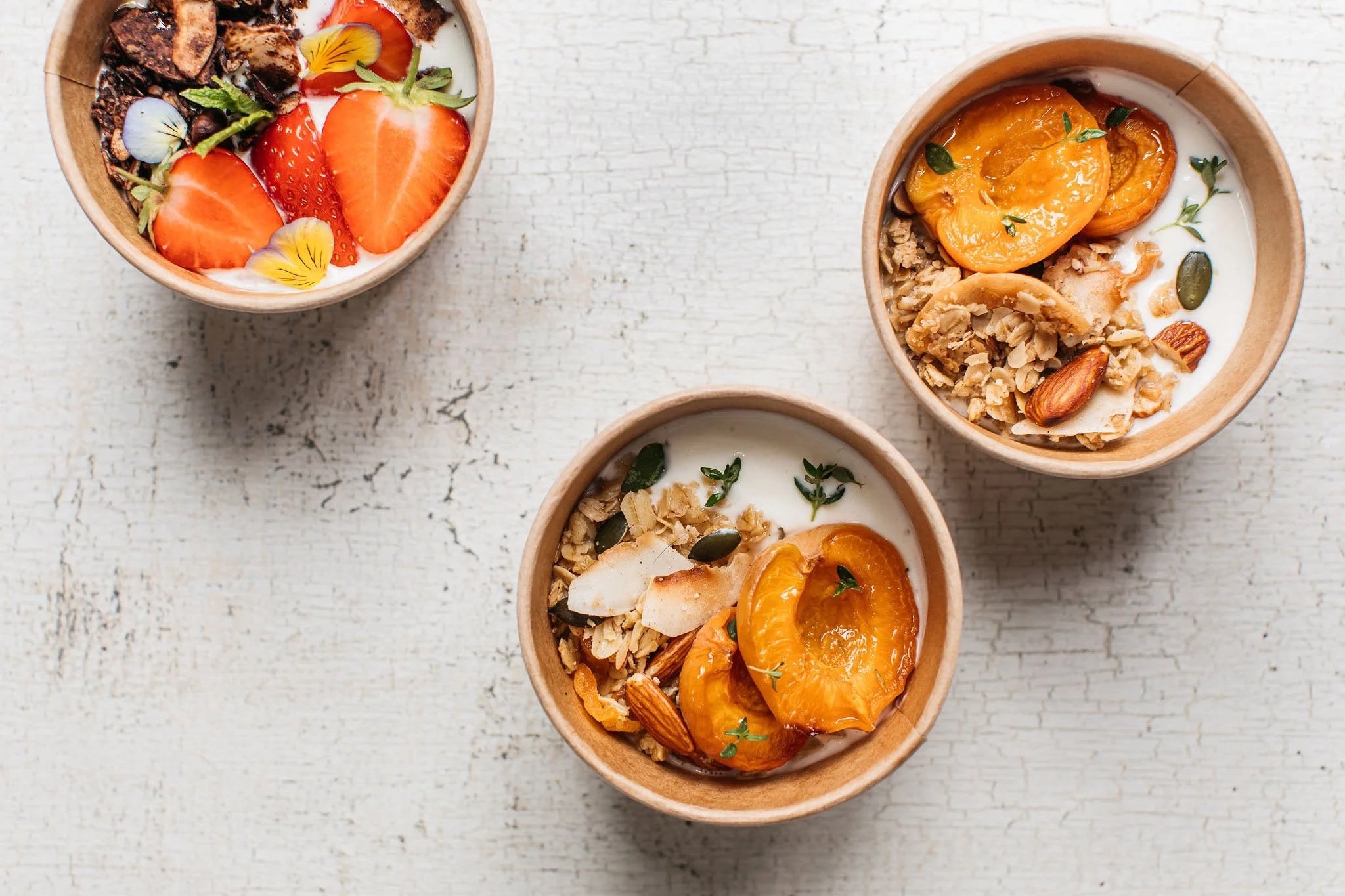 Three bowls containing yogurt with granola, sliced apricots, almonds, and herbs, topped with fruit slices and seeds, on a white textured surface.