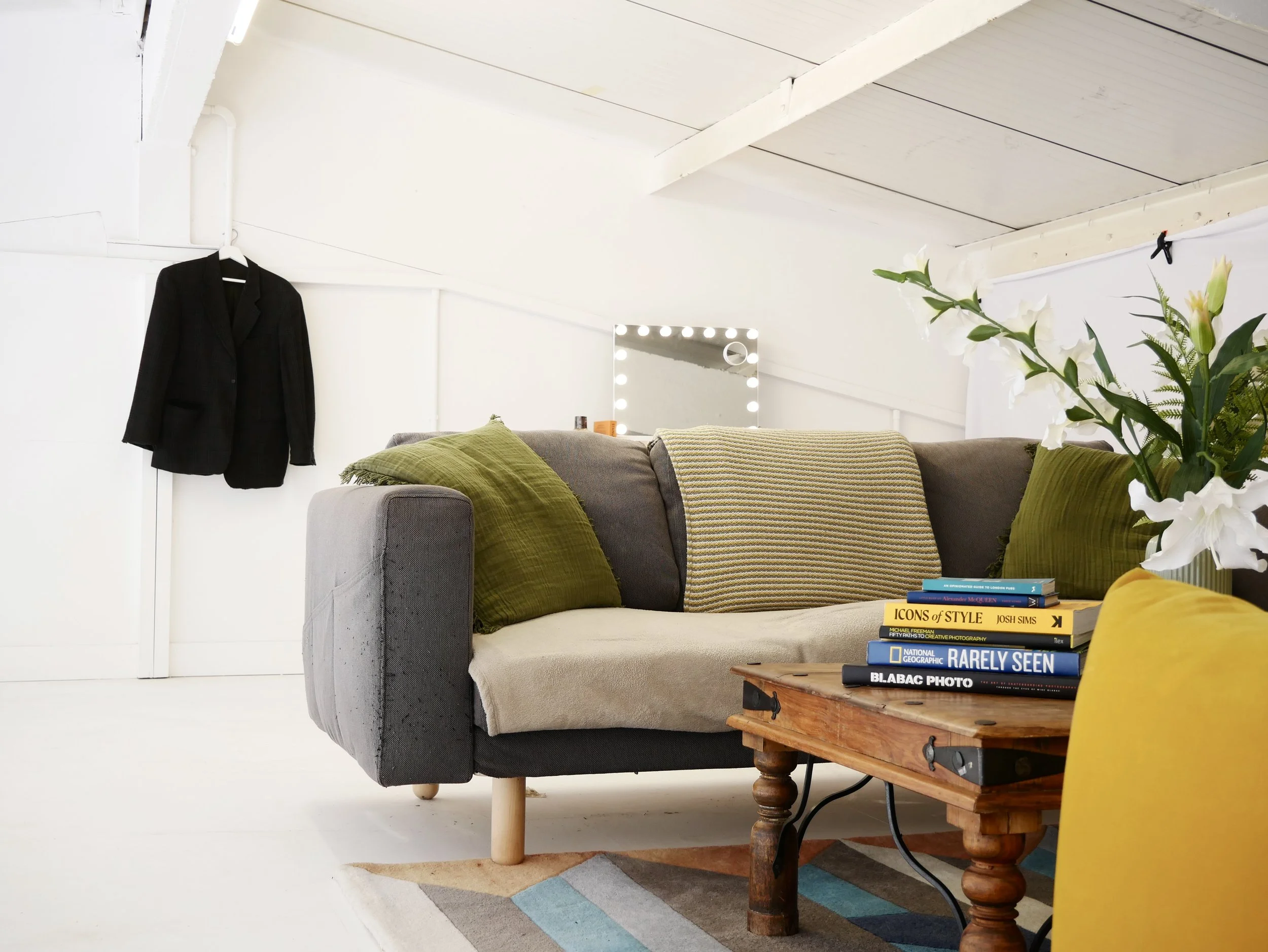 Living room with a gray couch with green and striped pillows, a wooden coffee table with books, a large vase with white lilies, a yellow chair, and a black blazer hanging on the wall. A vanity mirror with lights is in the background.