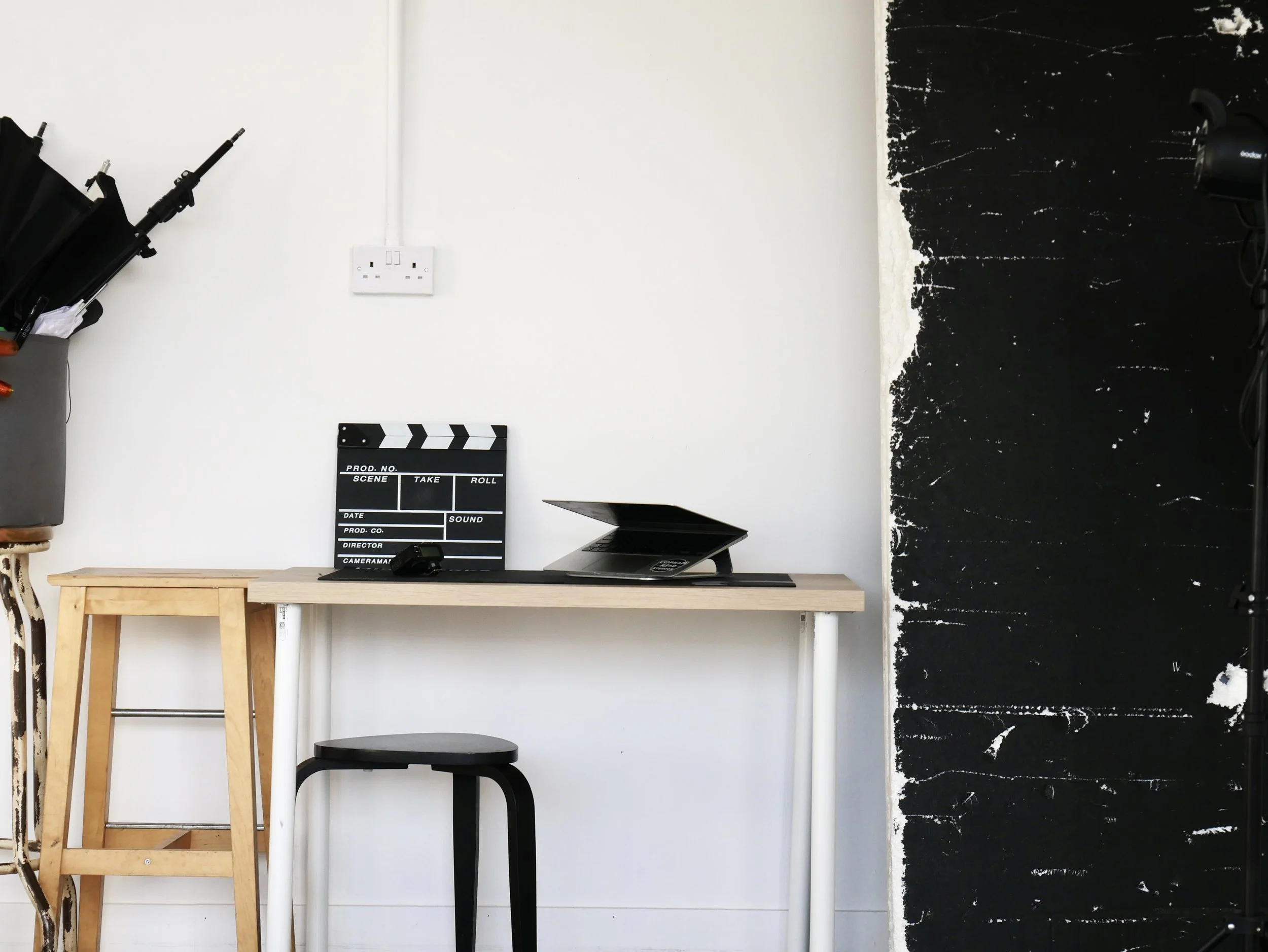 Interior of a studio or office with a wooden table, black stool, clapperboard, laptop, umbrella stand with umbrellas, and a black wall with some white paint splatters.