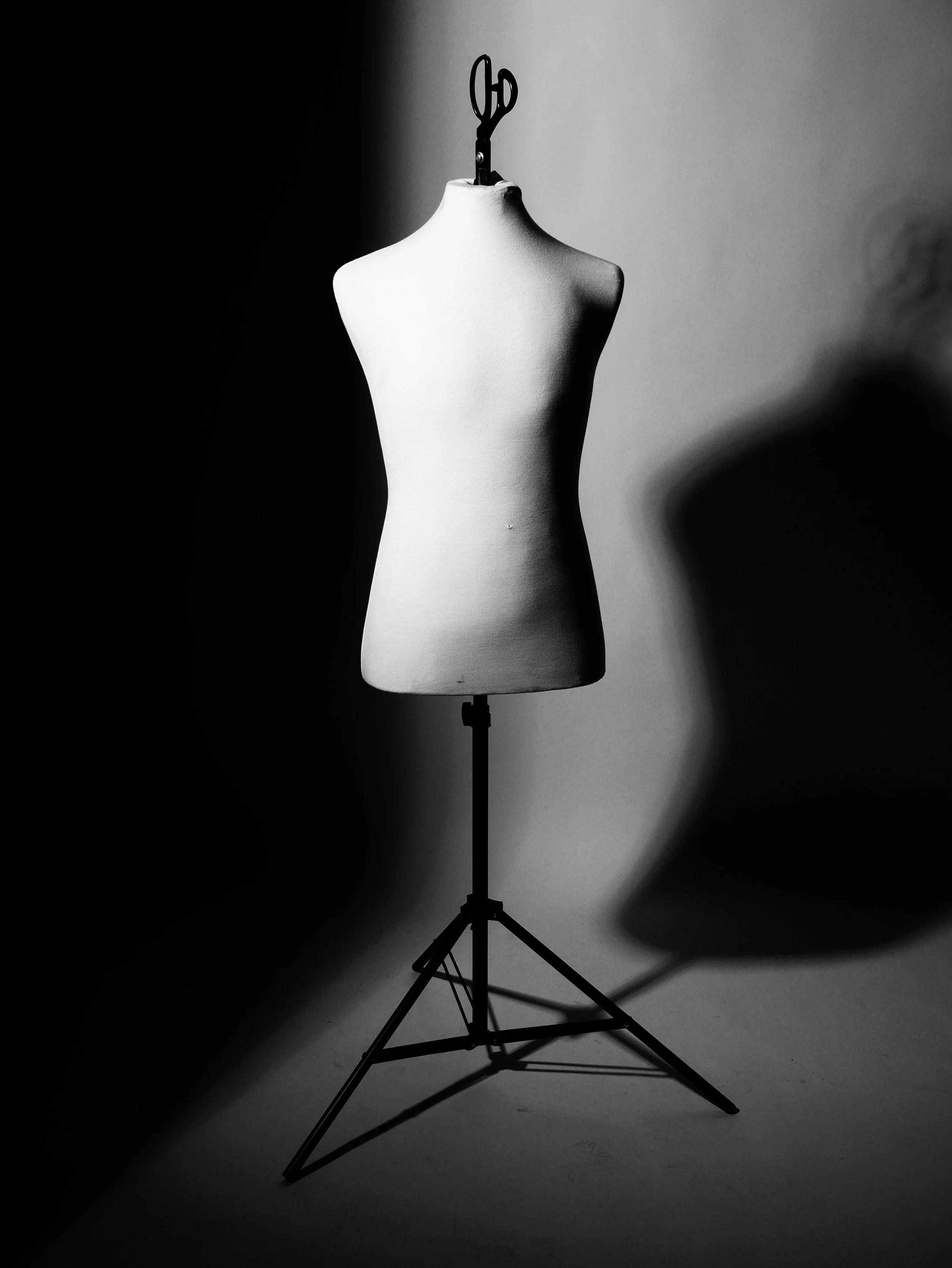 A black and white photograph of a dress form mannequin on a stand, casting a shadow on a plain background.