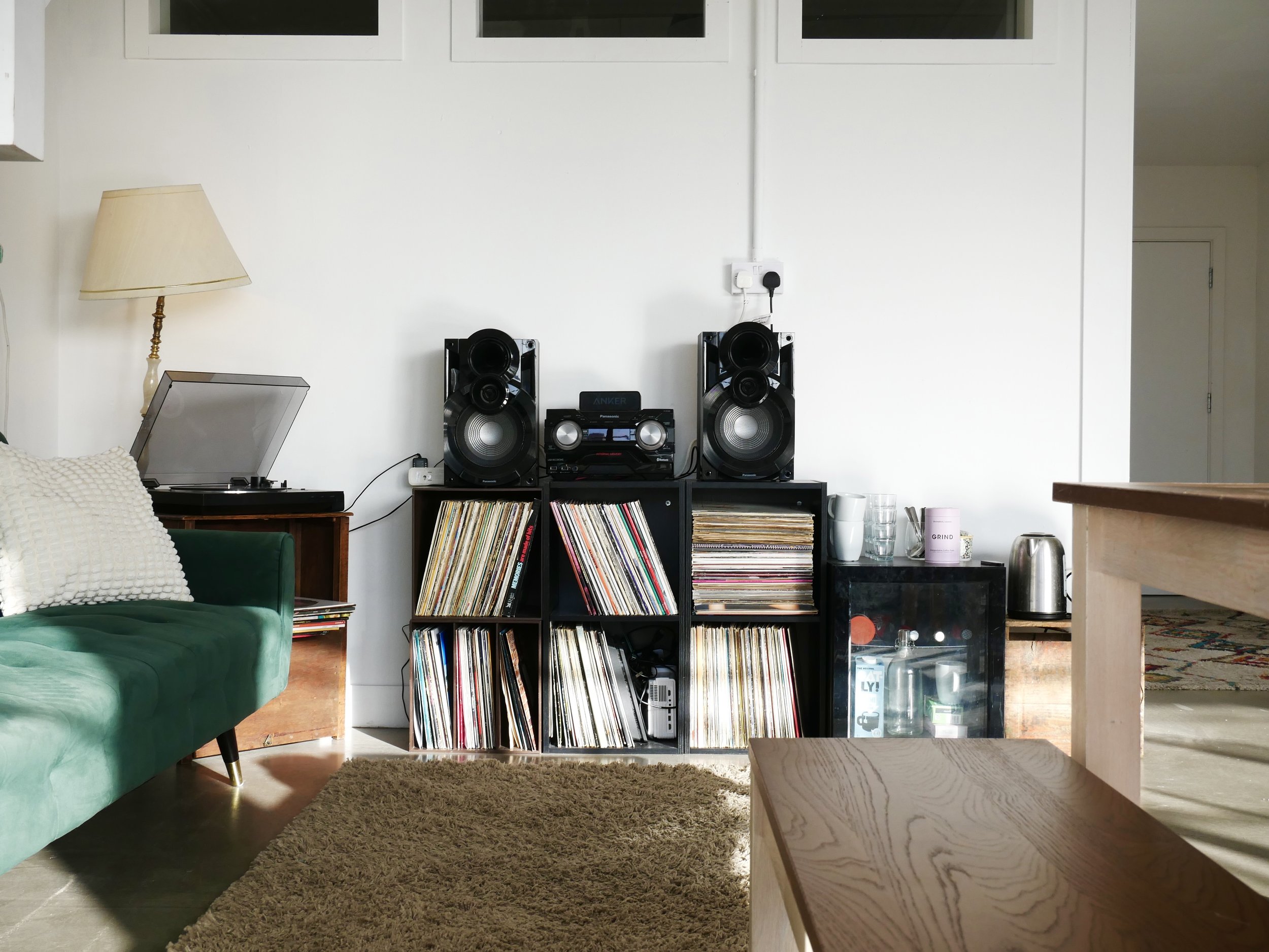 A living room with a green sofa, a beige carpet, a wooden side table, and a record player on a black shelf holding vinyl records. Two large black speakers and a stereo system are on top of the shelf. There is a lamp on a small wooden table to the lef