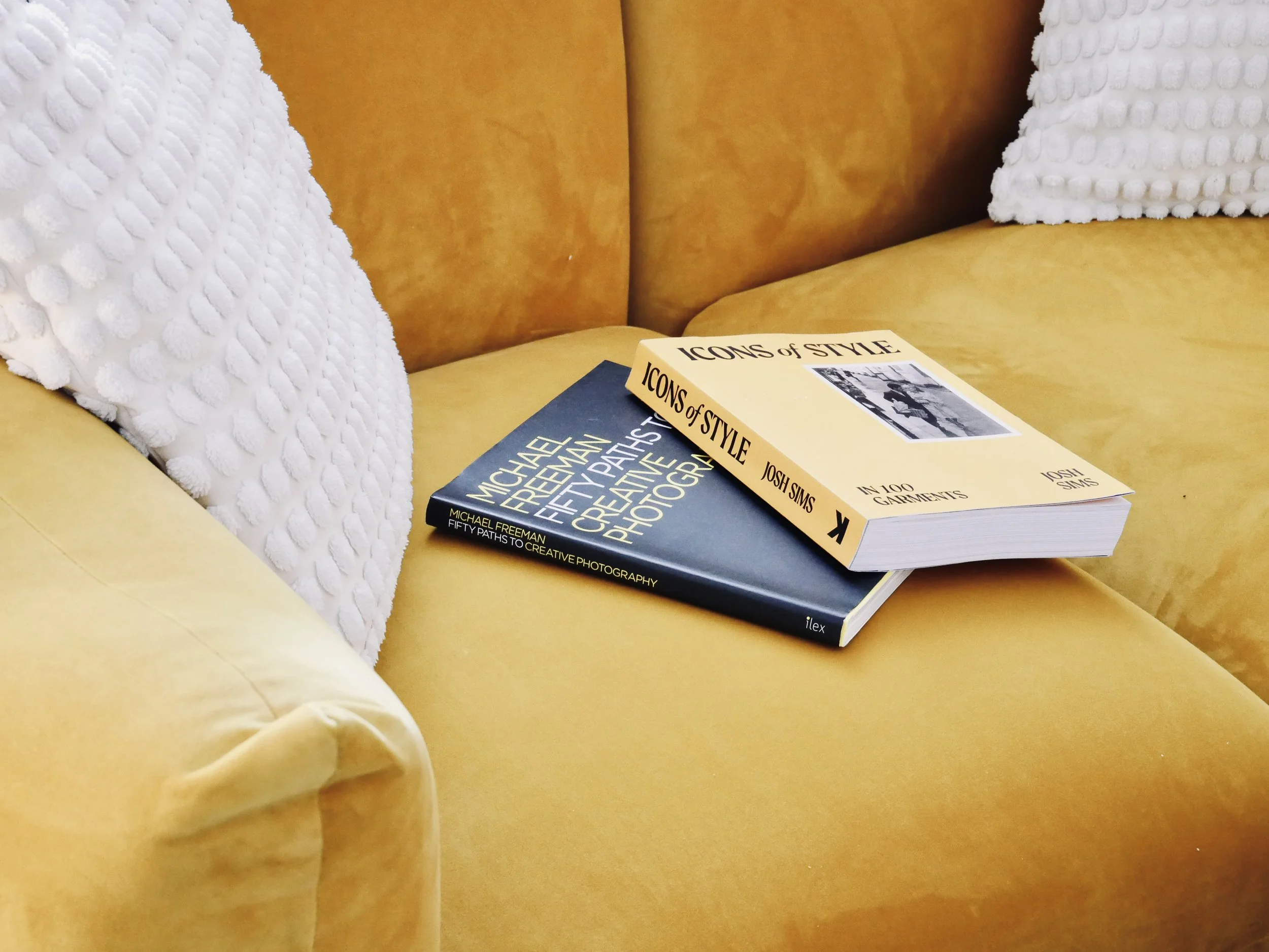 A yellow sofa with two white pillows and two books resting on the sofa, one titled 'Icons of Style' and the other 'Michael Freeman Fifty Paths to Creative Photography'.