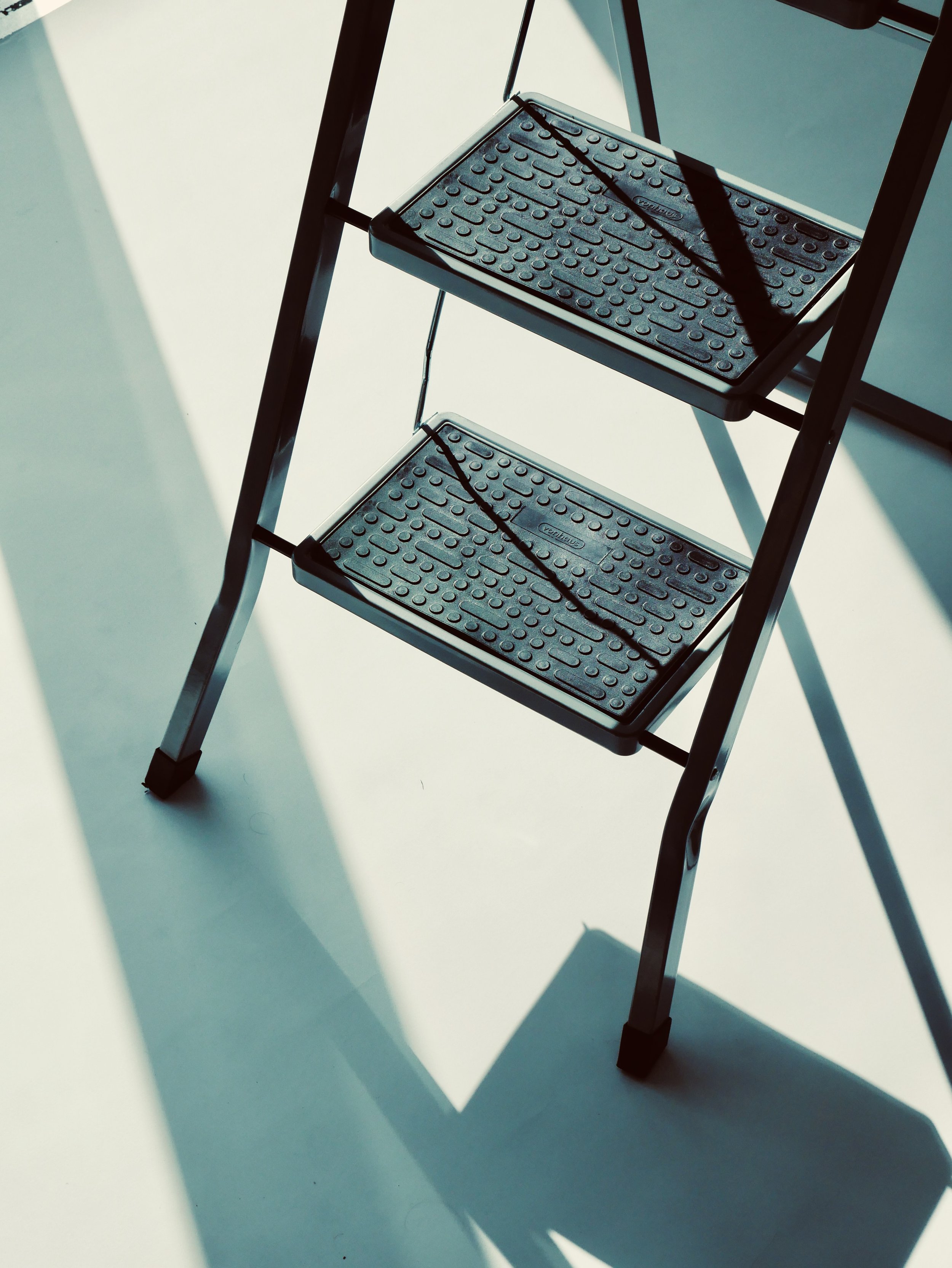 A black metal step ladder with two steps, each having a textured surface, standing on a smooth white floor with shadows cast by sunlight.
