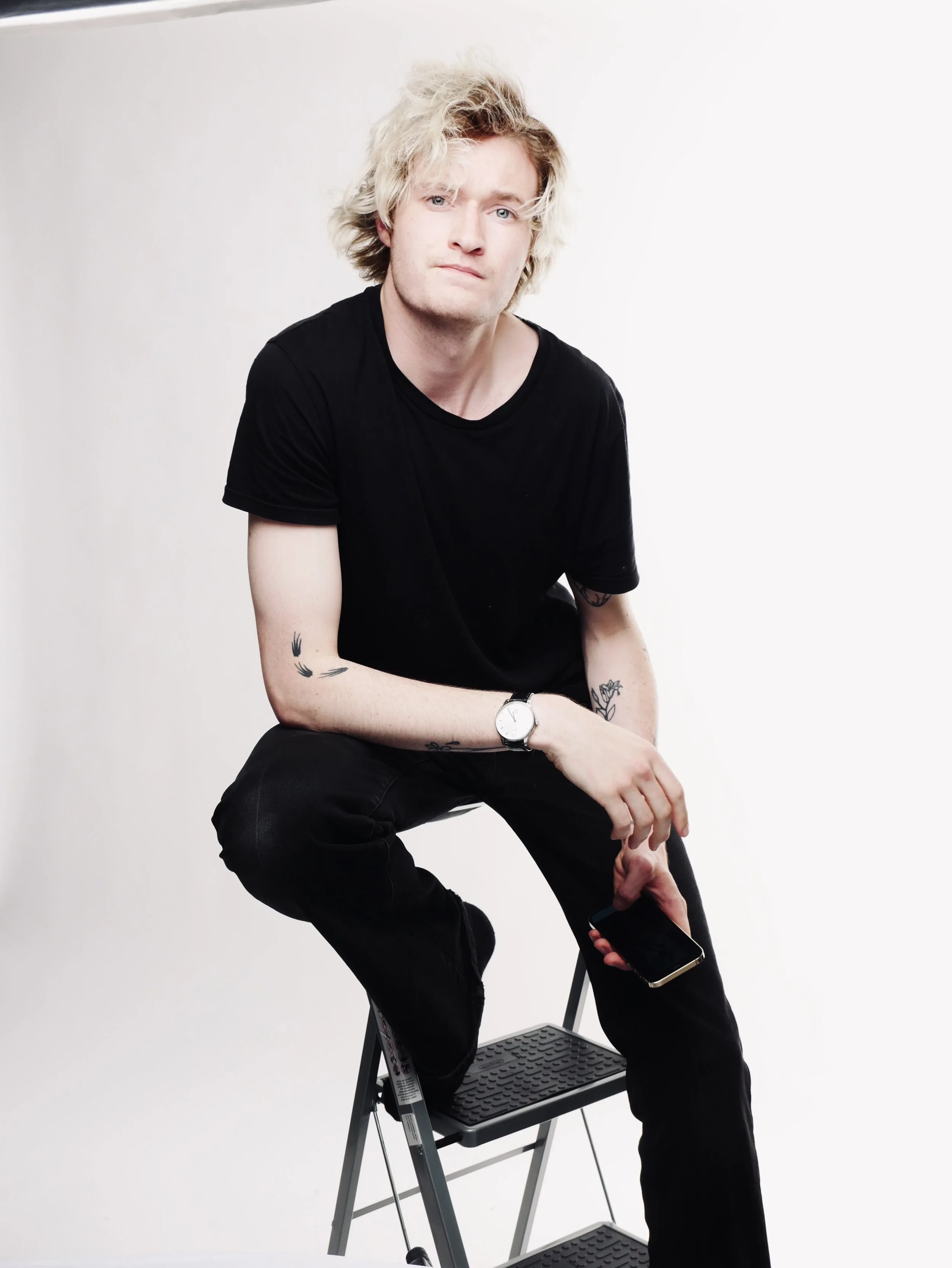 Young man with curly blonde hair, wearing a black t-shirt and black pants, sitting on a black step stool against a plain white background, holding a smartphone in his right hand, looking at the camera with a neutral expression.