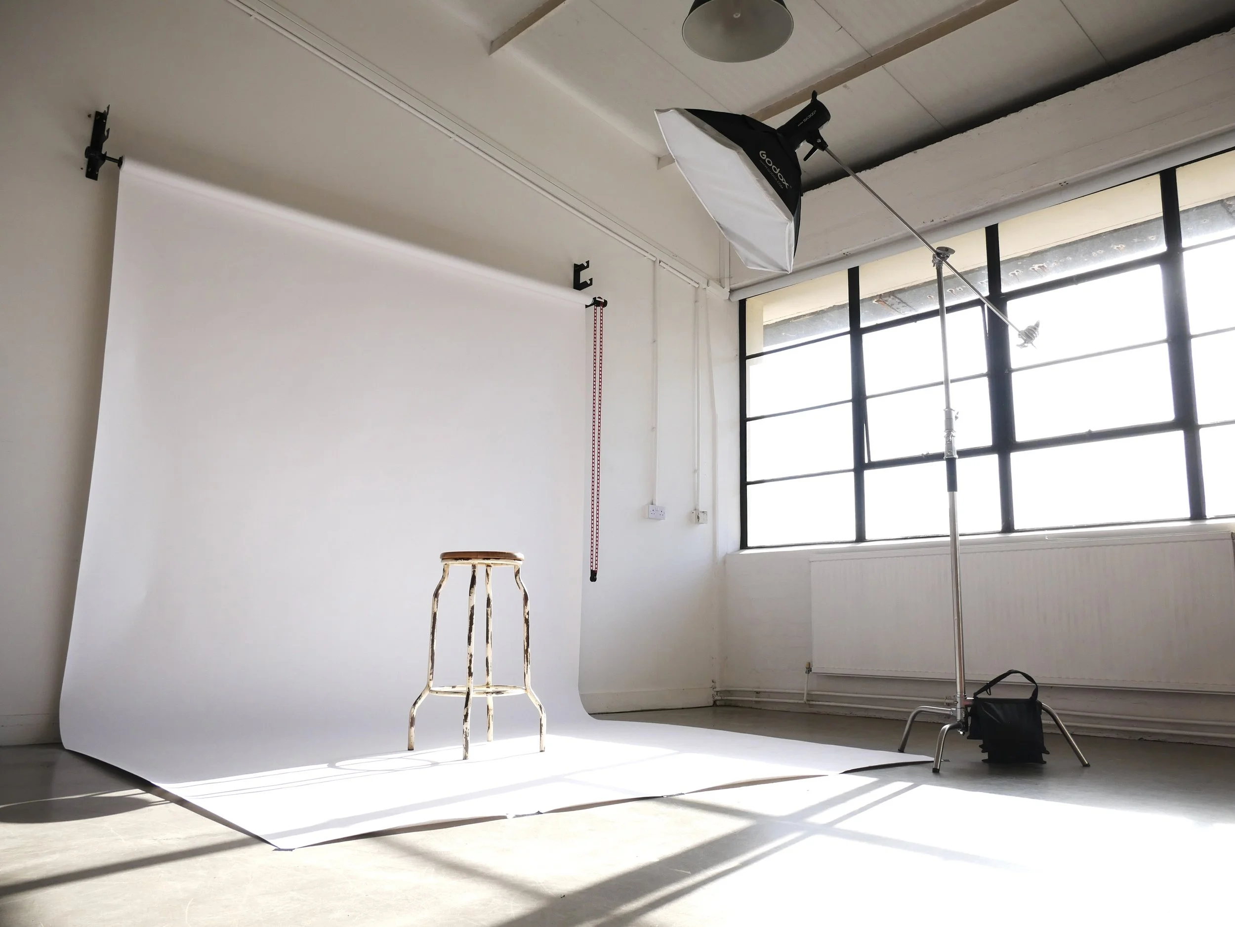 Photography studio with a white backdrop, a stool, and professional lighting equipment near a large window.