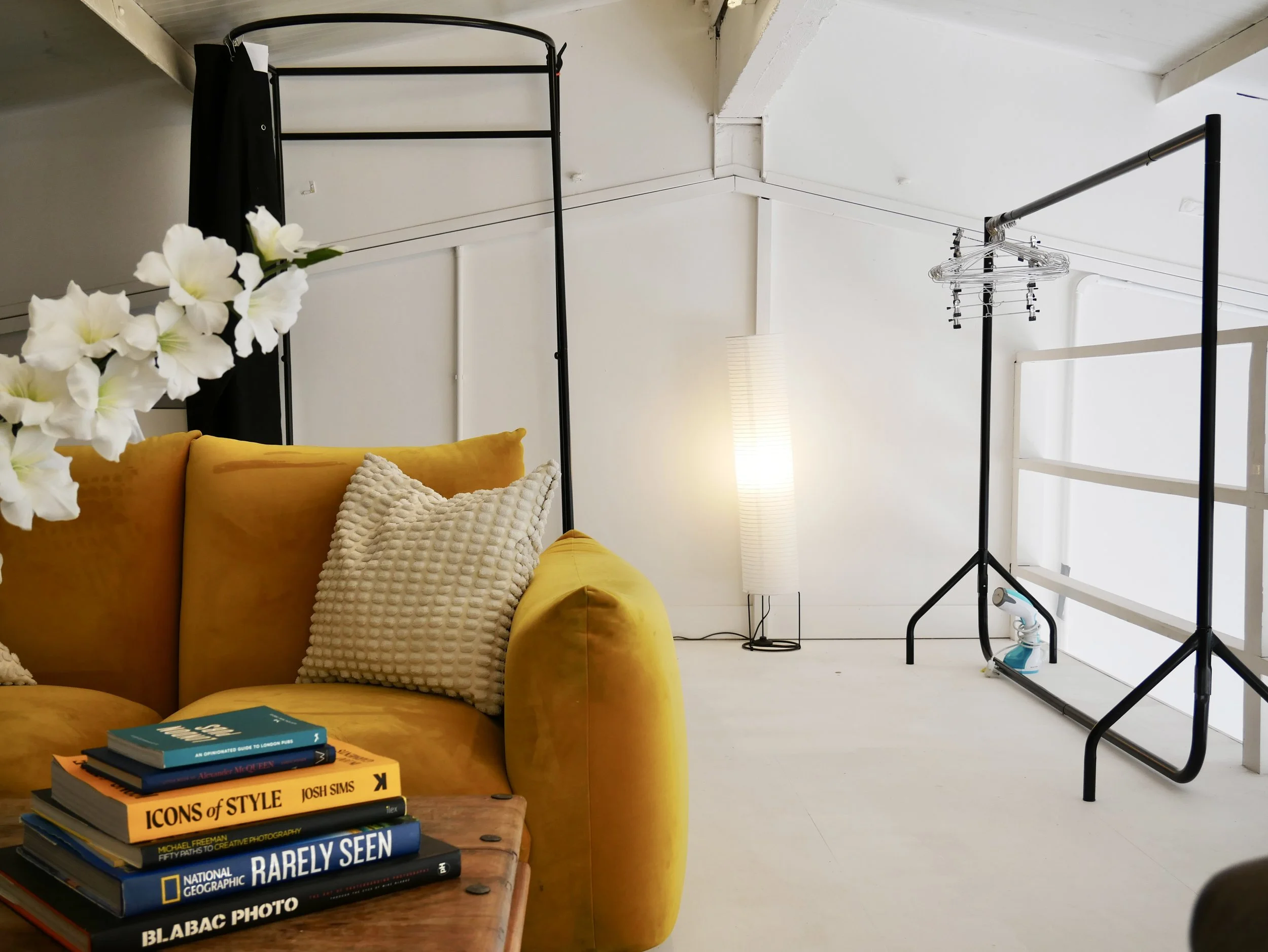 Interior of a room with a yellow velvet sofa, a stack of photography books on a wooden table, a white cushion, a standing floor lamp, and two clothing racks with hangers. White walls and a sloped ceiling.